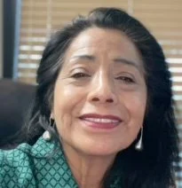Close-up of a woman with dark hair, smiling, wearing earrings and a green patterned top, seated in an indoor setting.