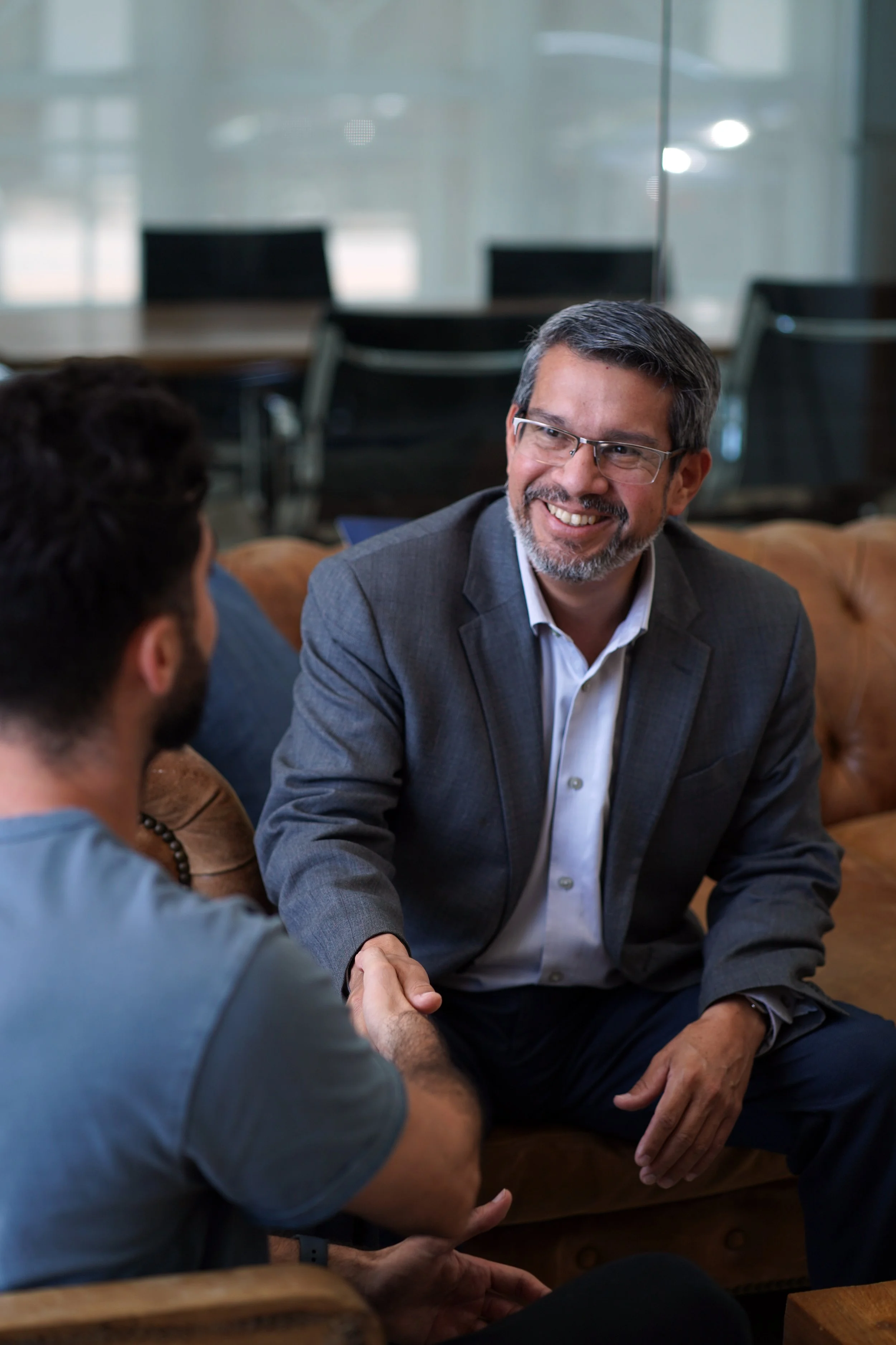 Two men shaking hands and engaging in conversation in a professional setting with a glass wall and multiple chairs in the background.