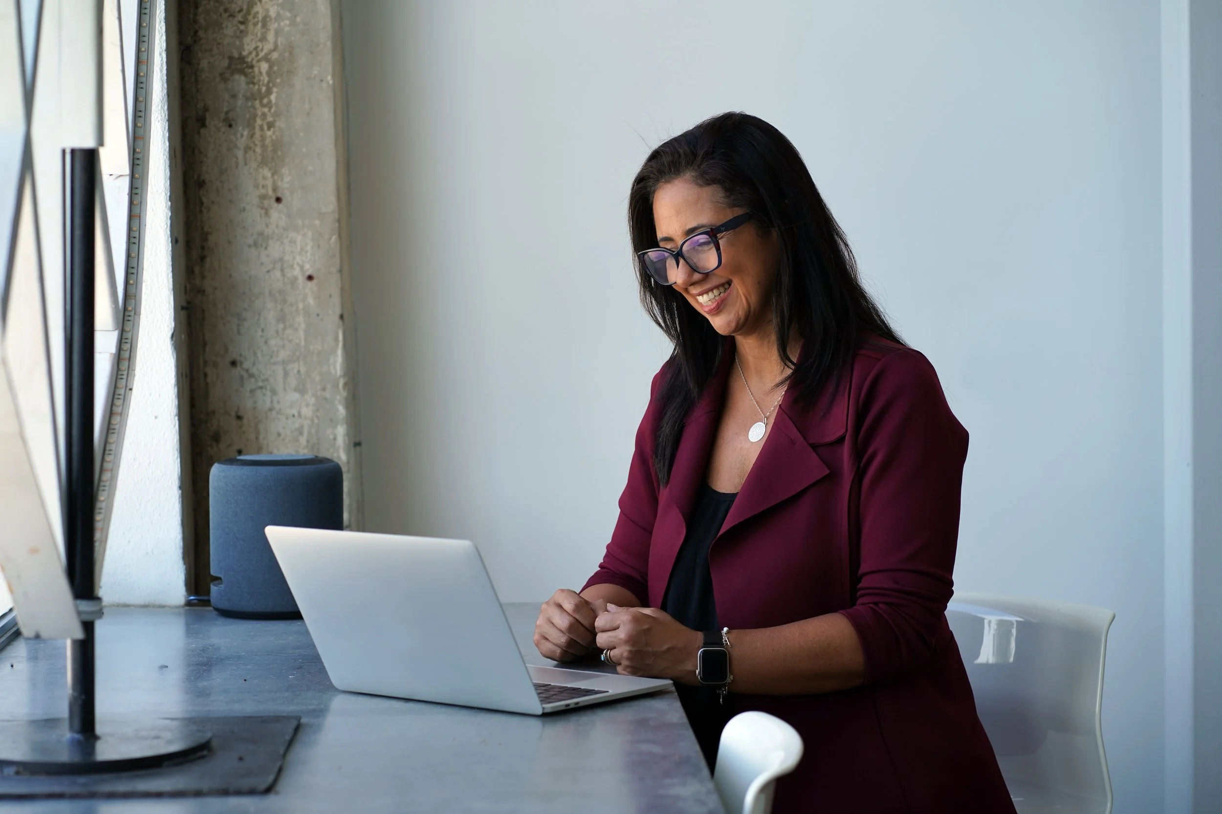 A woman with dark hair, glasses, and a burgundy blazer working on a laptop at a desk in a modern office setting, smiling and looking down.