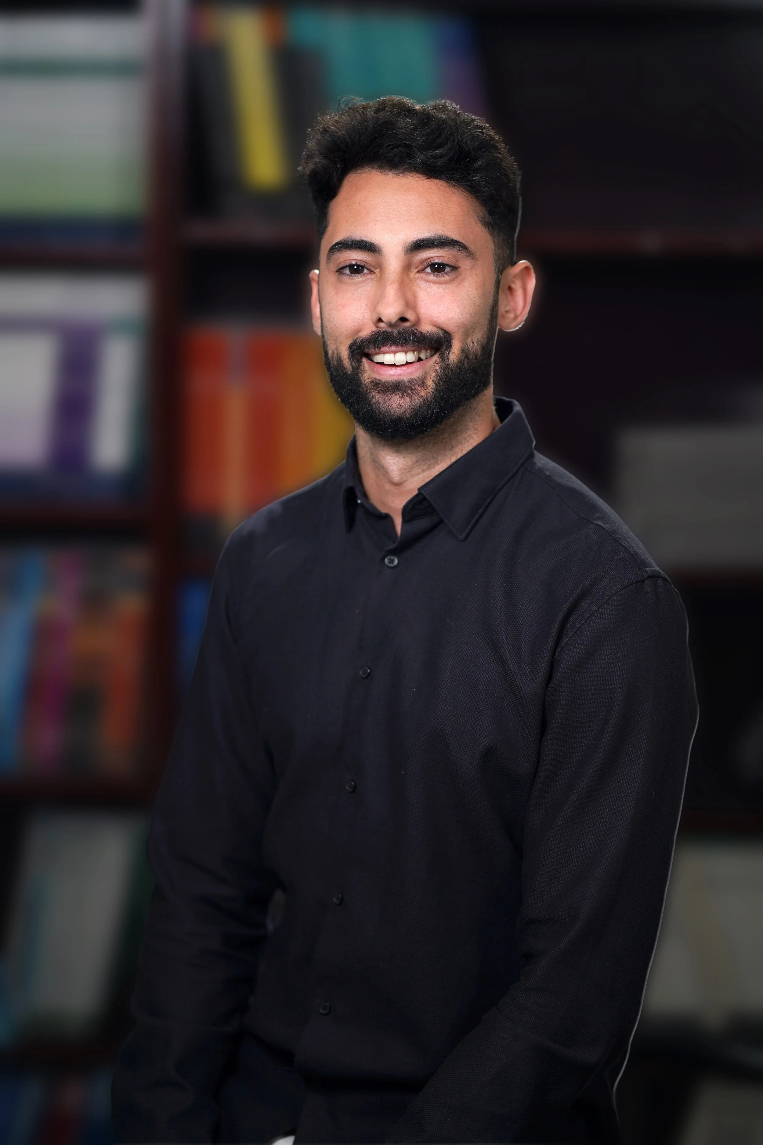 A young man with dark hair and a beard, wearing a black button-up shirt, smiling in front of a blurred bookshelf background.