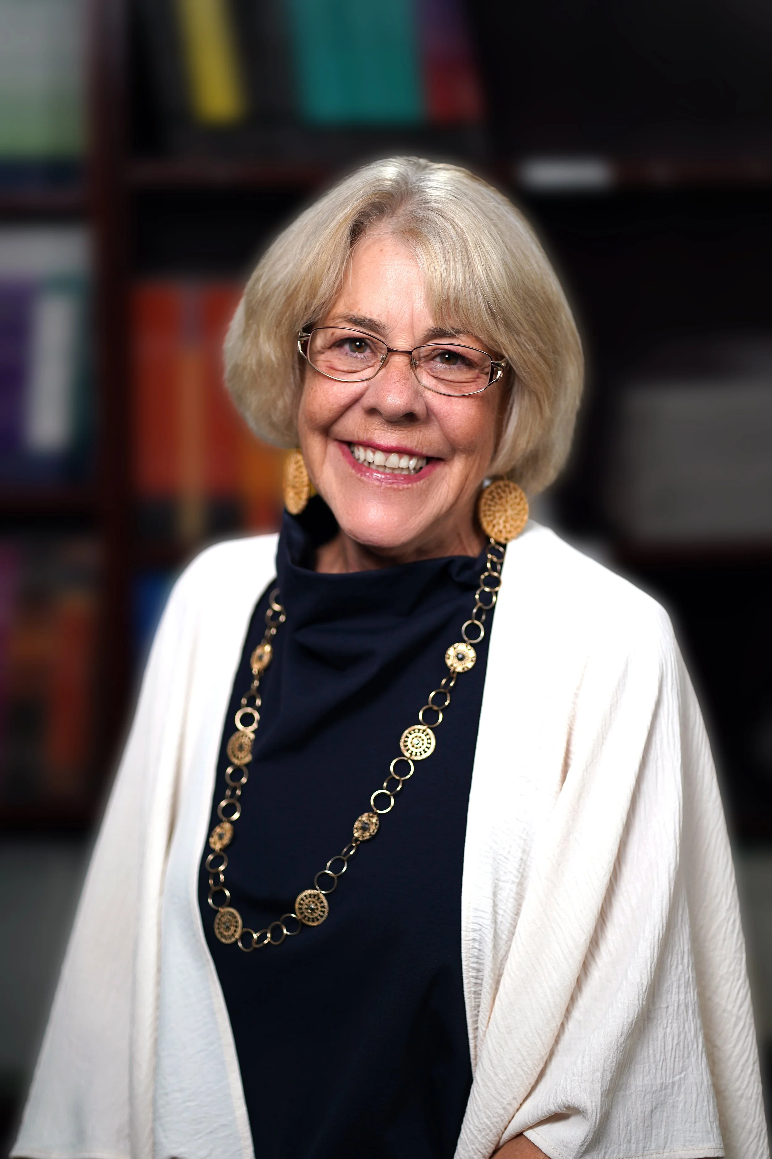 An older woman with blonde hair and glasses smiling, wearing a black top, white cardigan, and gold jewelry, standing in front of a blurred bookshelf.