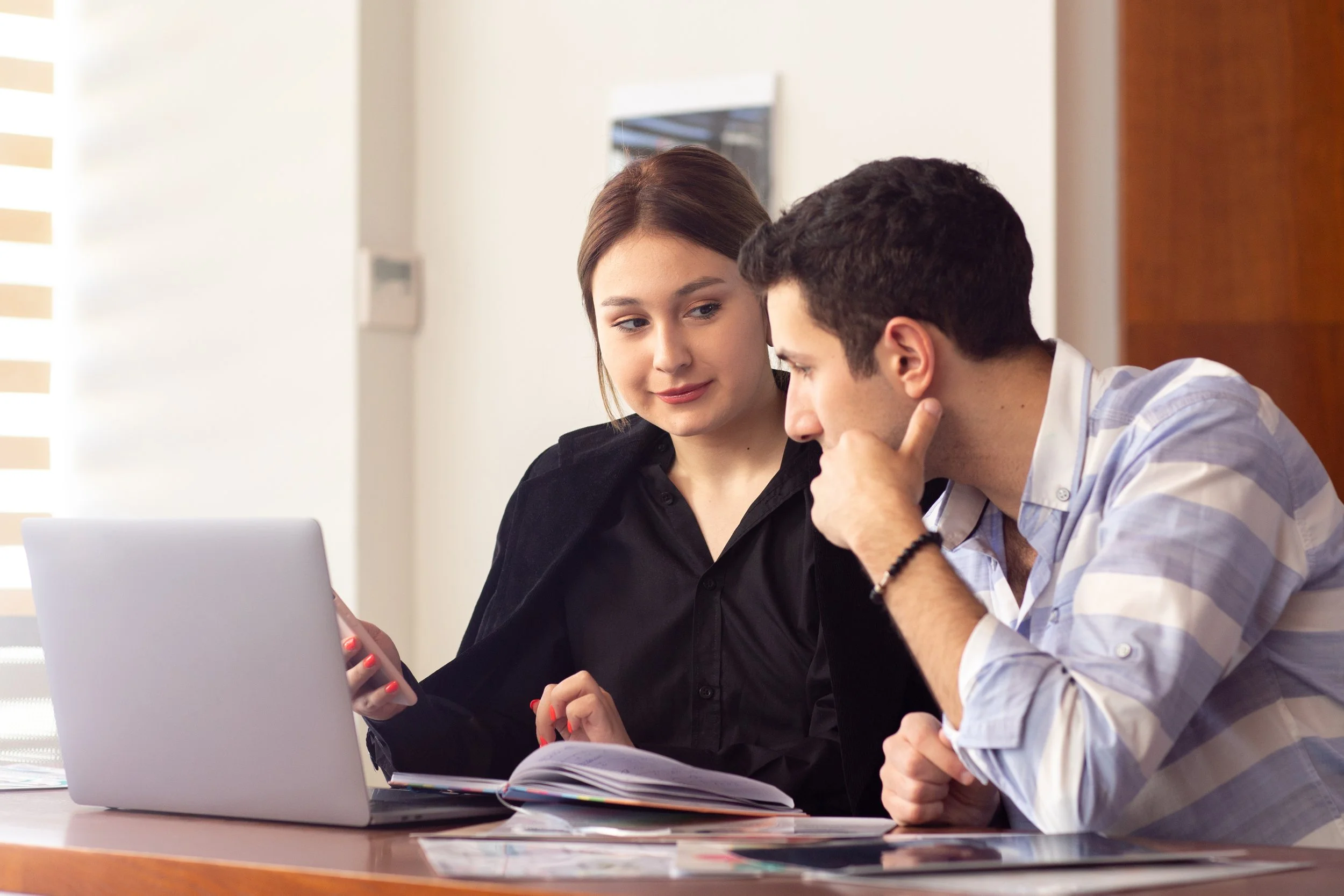 A young woman and a young man sit at a desk, looking at a laptop screen and a notebook, engaged in a discussion.