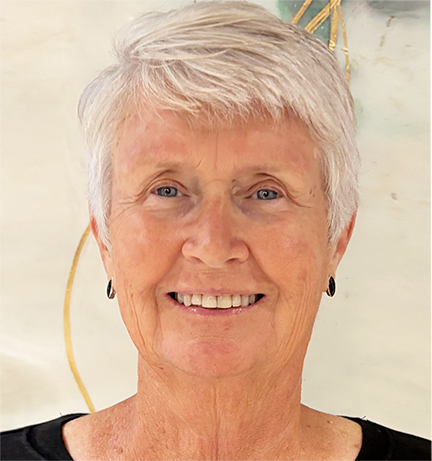 A smiling older woman with short, gray hair and black earrings, wearing a black top, standing in front of a light-colored background.