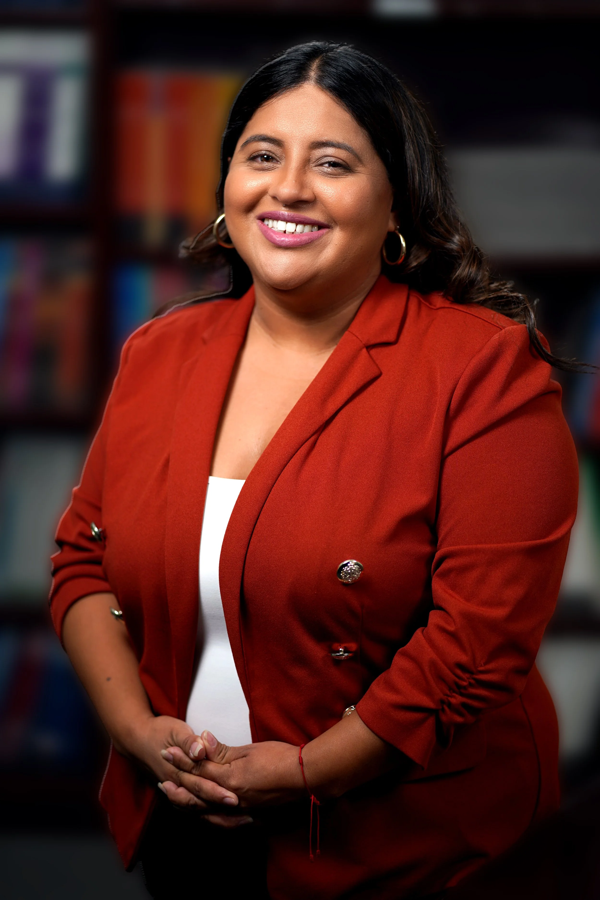 A woman smiling, wearing a red blazer and hoop earrings, standing in front of a blurred background of bookshelves.