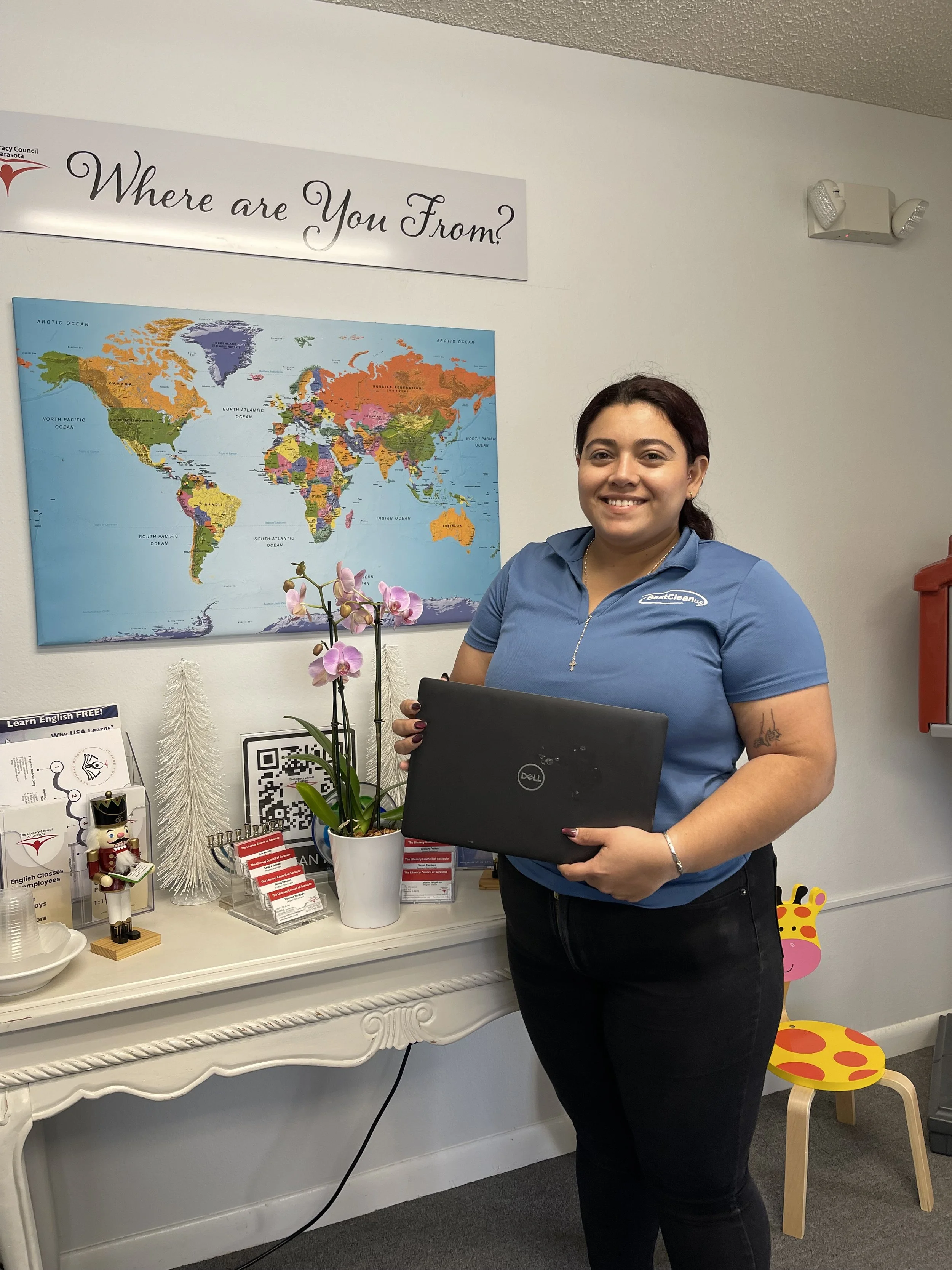 A woman with dark red hair, wearing a blue polo shirt, holding a black Dell laptop, standing in front of a white desk with various decorative items, including a pink orchid plant, a nutcracker figure, and informational brochures. Behind her is a world map on the wall, a sign that says 'Where are You From?' and a small colorful chair shaped like a giraffe.