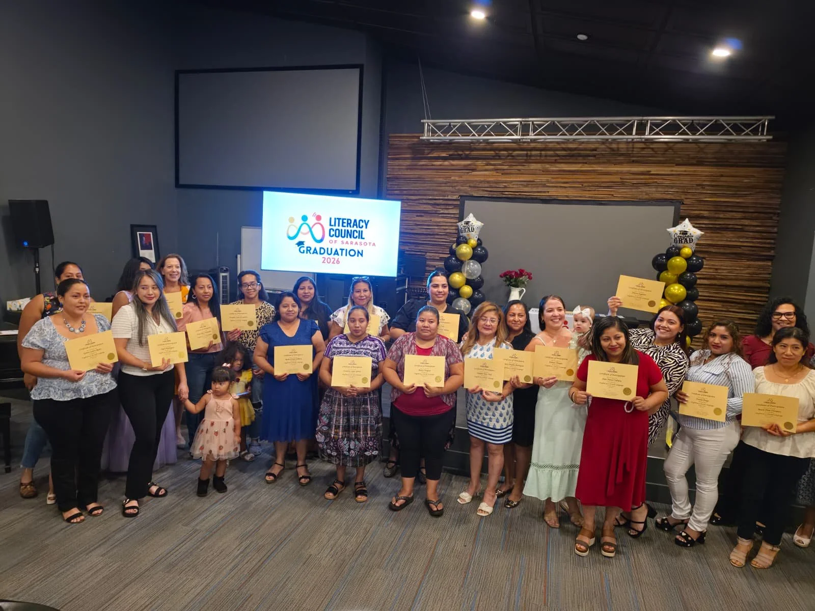 Group of adults and children at a graduation ceremony holding diplomas, with a screen showing 'Literacy Council of Sarasota Graduation 2026' in the background.
