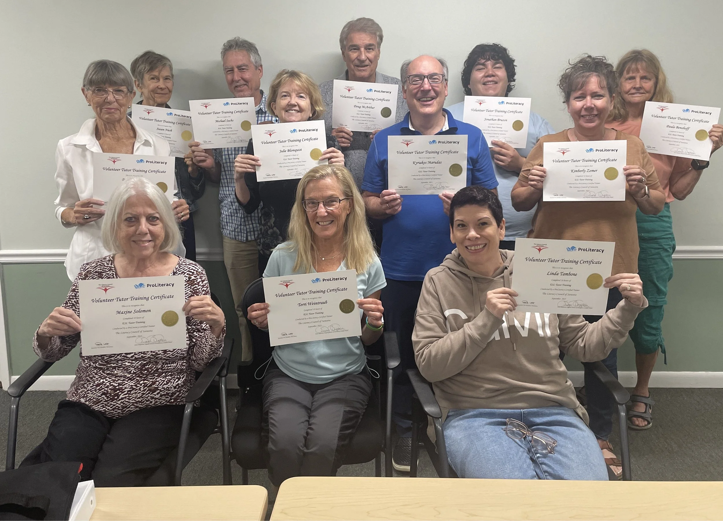 A group of diverse older adults holding certificates for volunteer tutor training, smiling, at an indoor event.