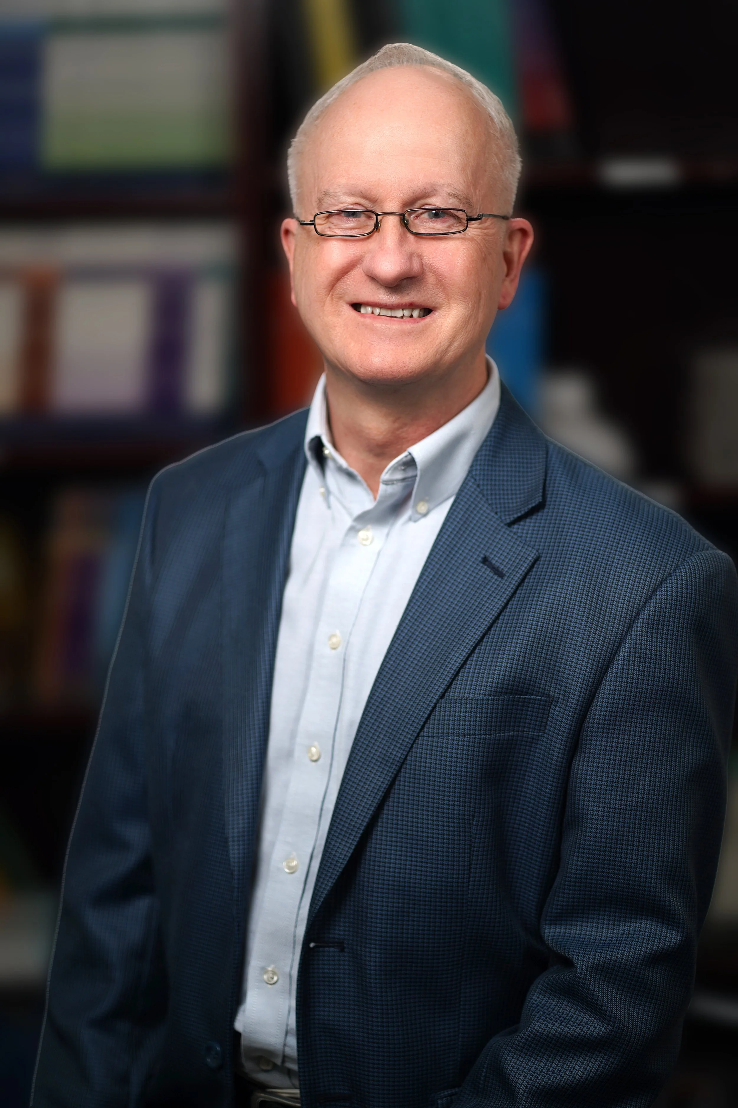 Portrait of a smiling middle-aged man with glasses, wearing a navy blue blazer and a white shirt, in front of blurred bookshelves.