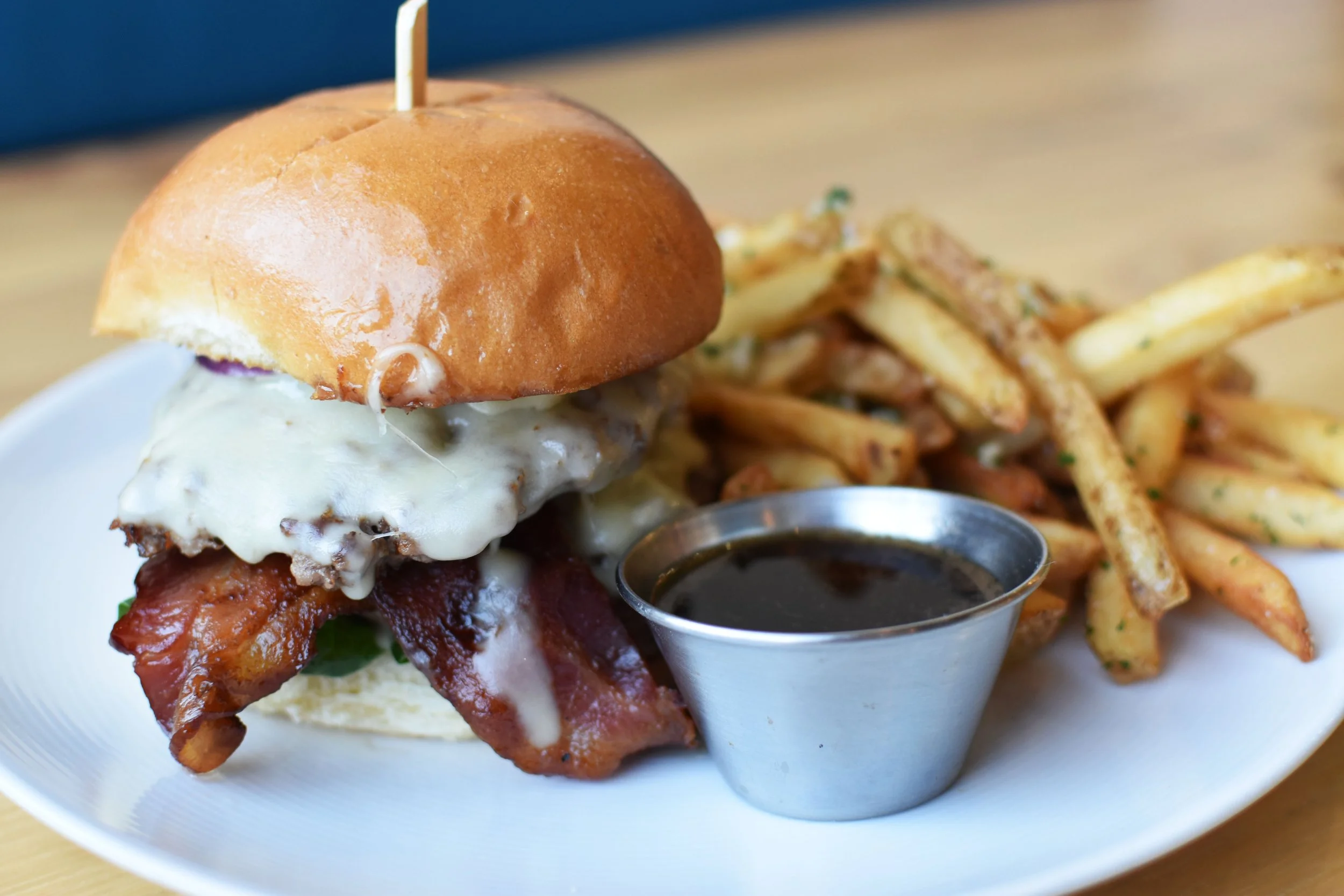 Burger with cheese, bacon, lettuce, and a bun served with French fries and a cup of dark dipping sauce