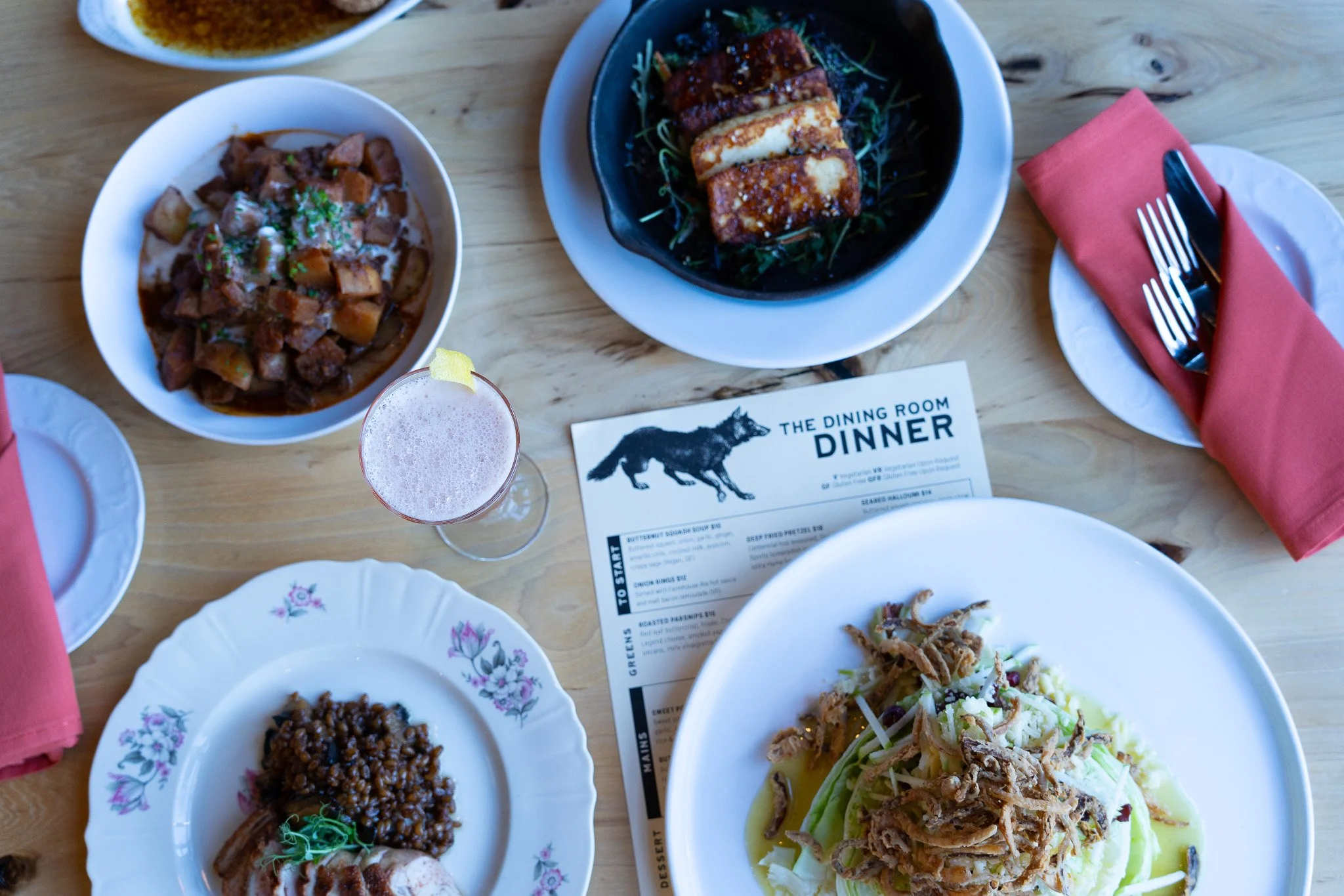 A table set with various dishes including a bowl of beef stew, a plate with sliced roasted meat, a plate with lentils and another with shredded meat on vegetables, a drink with a lemon twist, silverware on a pink napkin, and a dinner menu with a wolf graphic.