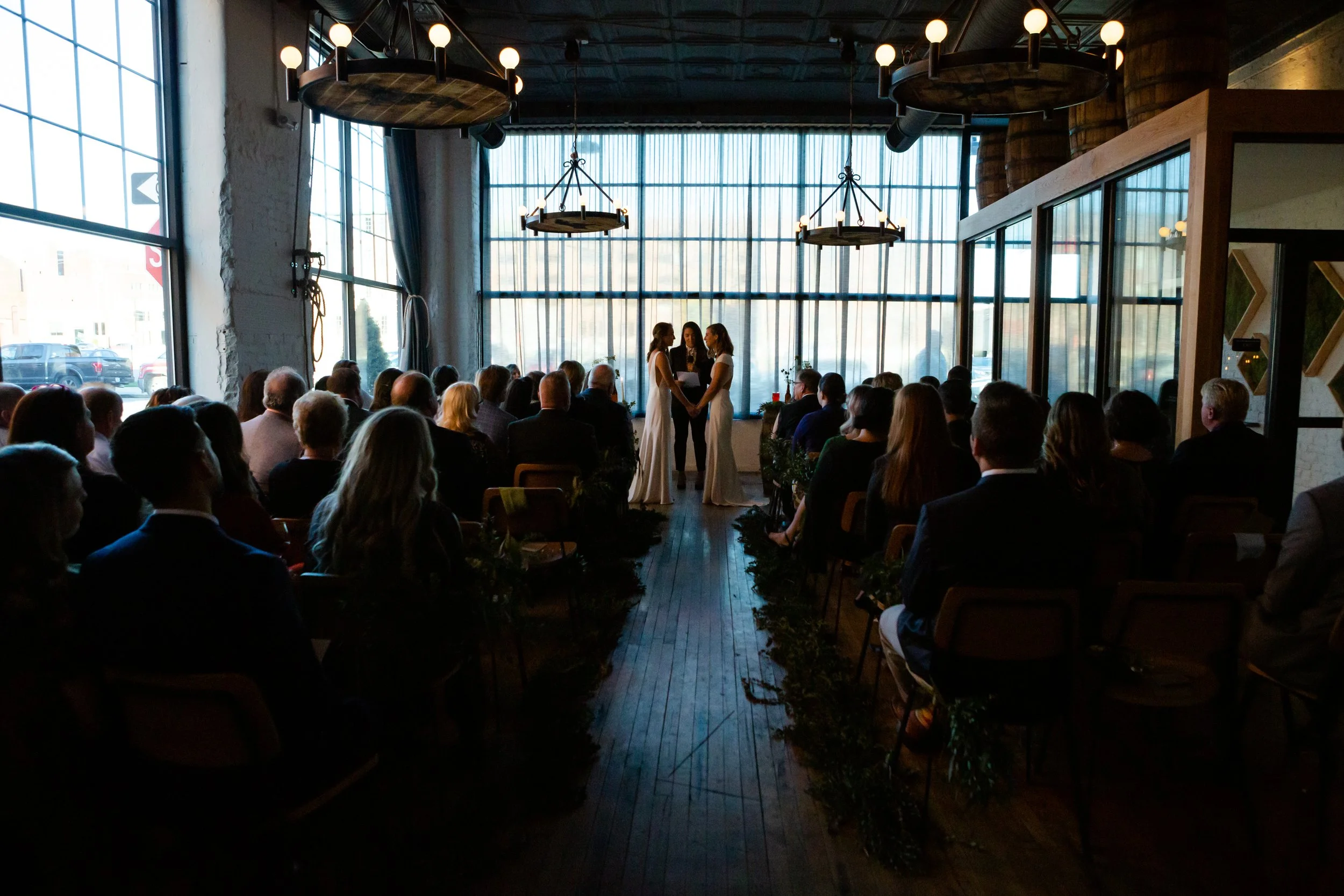 Indoor wedding ceremony with three women standing and holding hands at the altar, guests seated facing them, large windows in the background, natural light filling the room, overhead chandeliers, wooden floor, and modern decor.