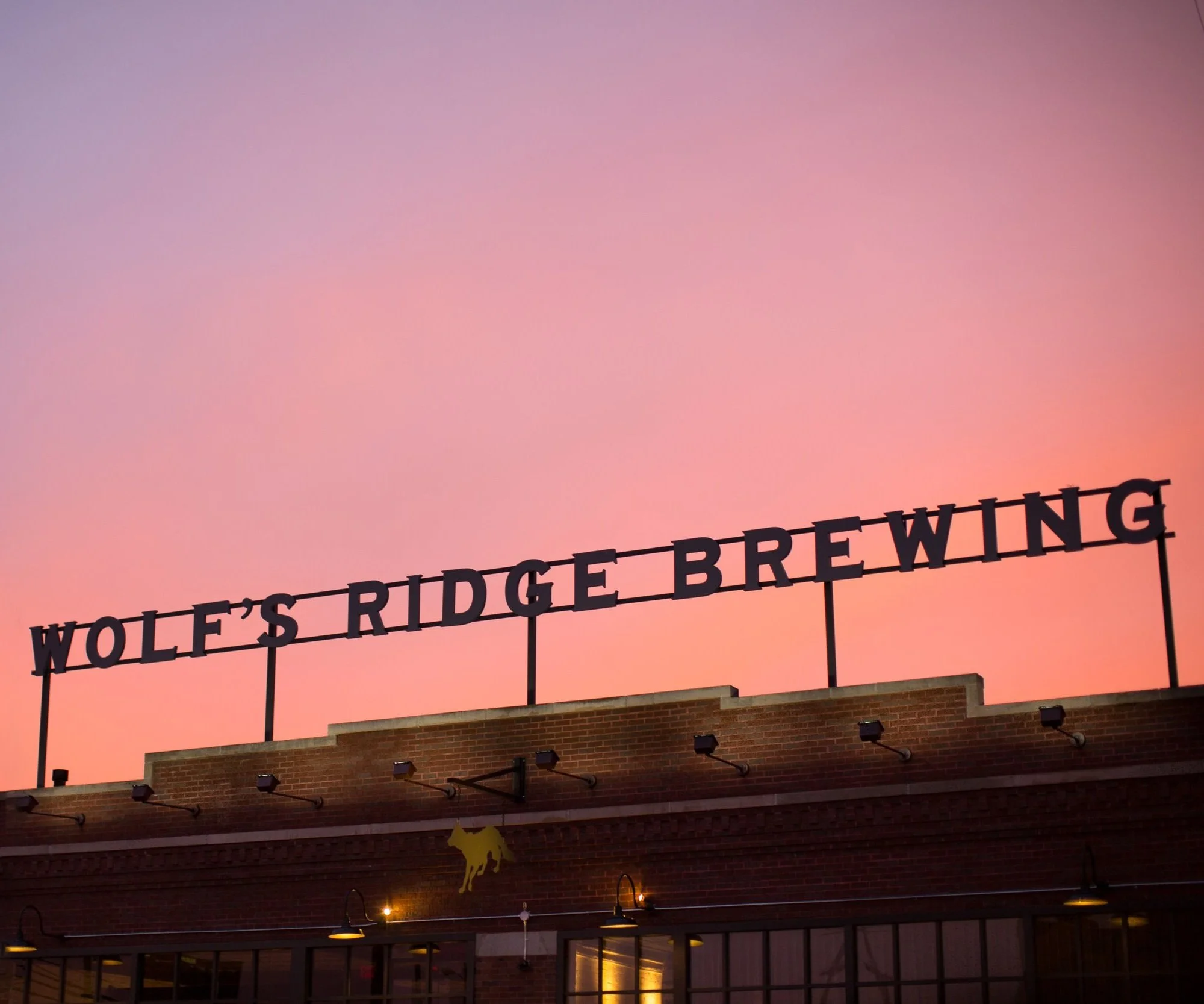 The sign 'WOLF'S RIDGE BREWING' on a brick building with a sunset sky in the background.