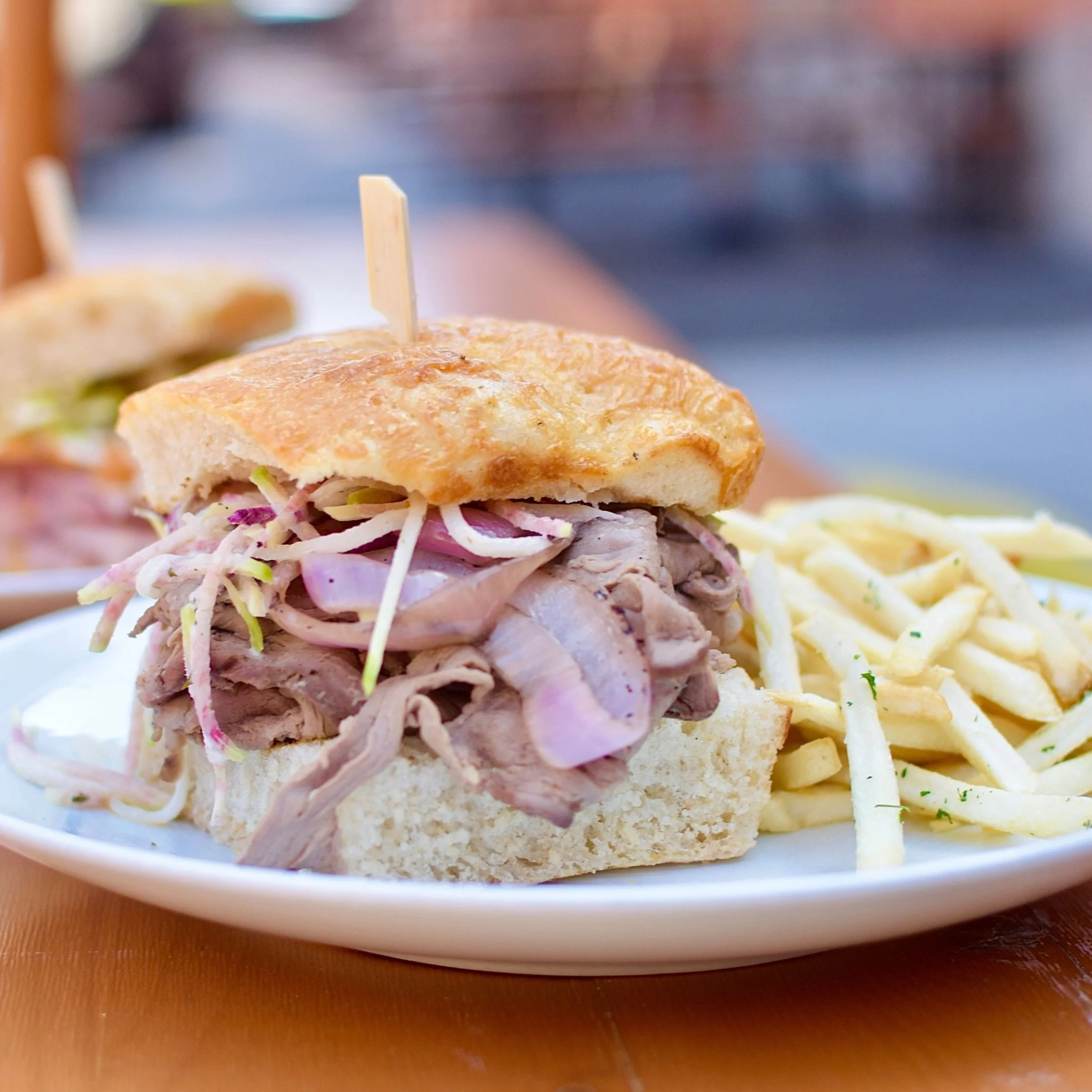 Roast beef sandwich with onions and coleslaw on a bun, served with French fries on a white plate.