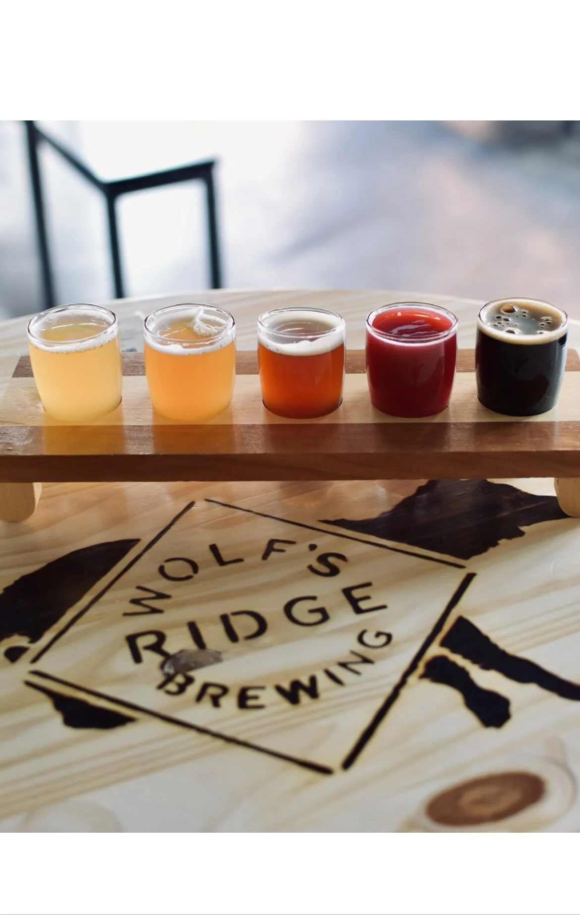 A flight of five small glasses of beer on a wooden tray, placed on a table with 'Wolf's Ridge Brewing' logo.