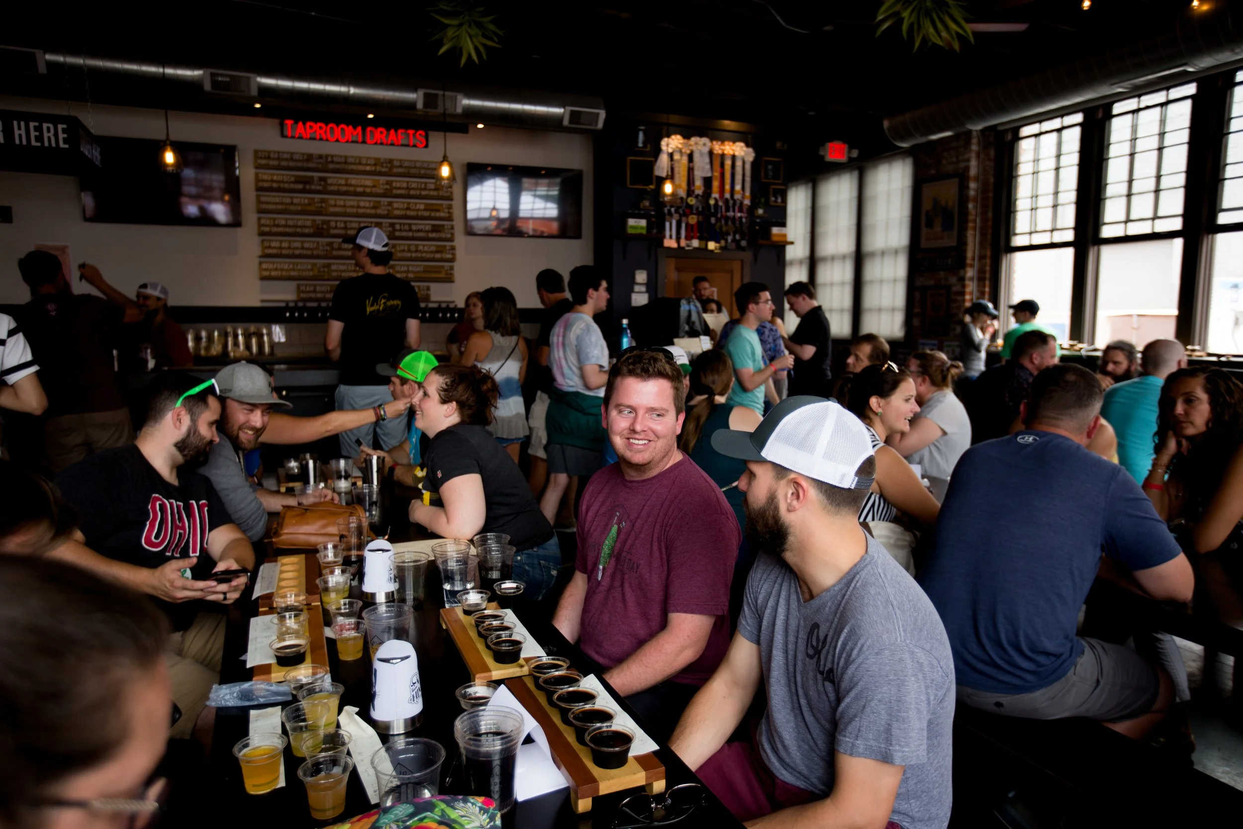 Group of people participating in a beer tasting event inside a brewery, sitting at a long table with several samples of beer in front of them, smiling and talking.