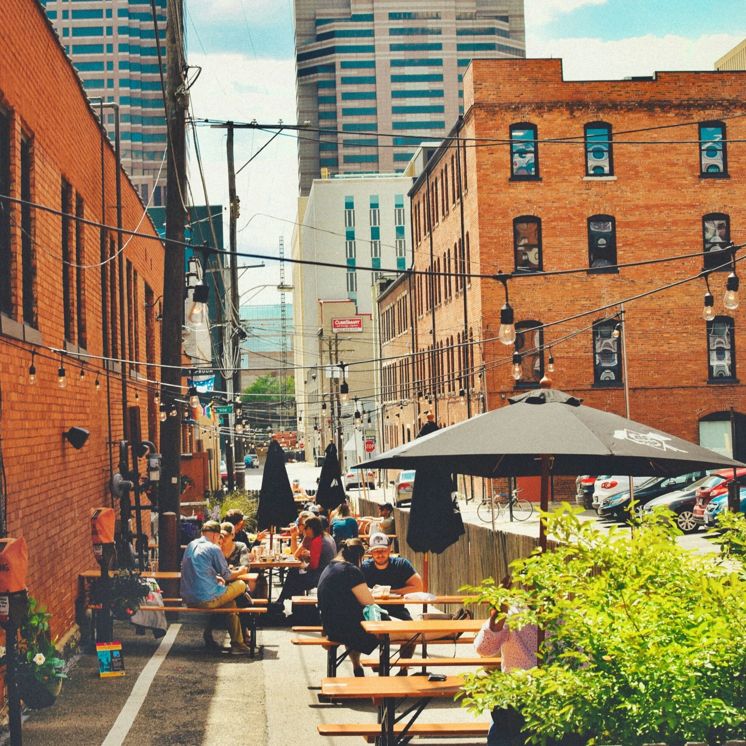 Outdoor dining area with people sitting at wooden tables, shaded by large umbrellas, on a city street with brick buildings, string lights, and tall modern skyscrapers in the background.