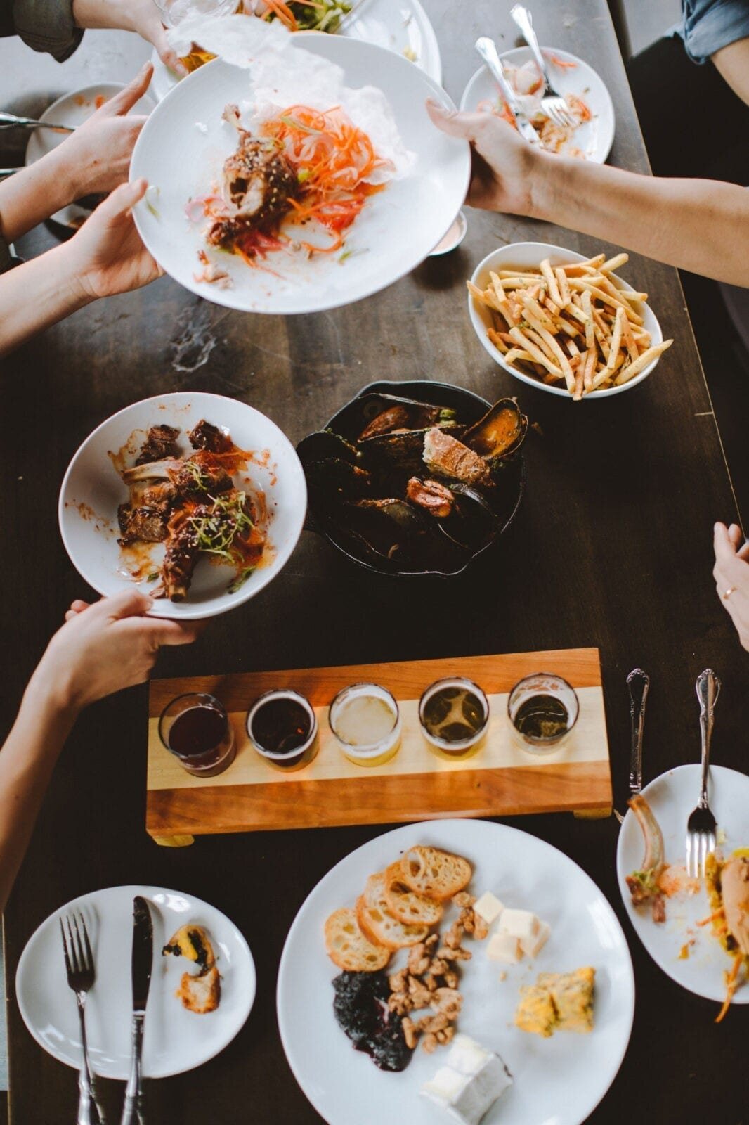 People sharing a meal with various dishes including rice, vegetables, sauces, seafood, bread, and cheese, along with a flight of five different beverages.
