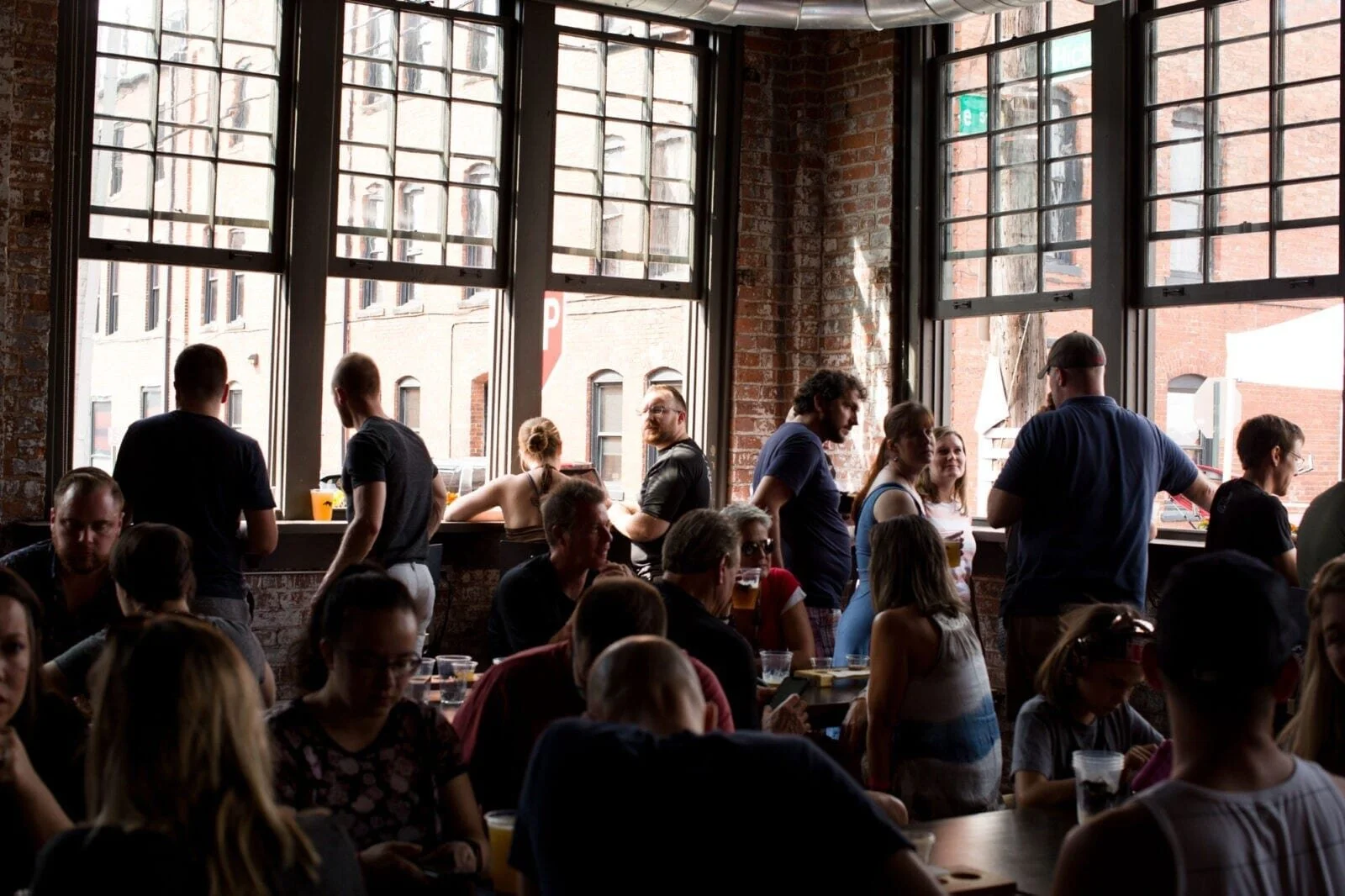 Interior of a busy brewery or bar with large windows, exposed brick walls, and people socializing and ordering drinks.