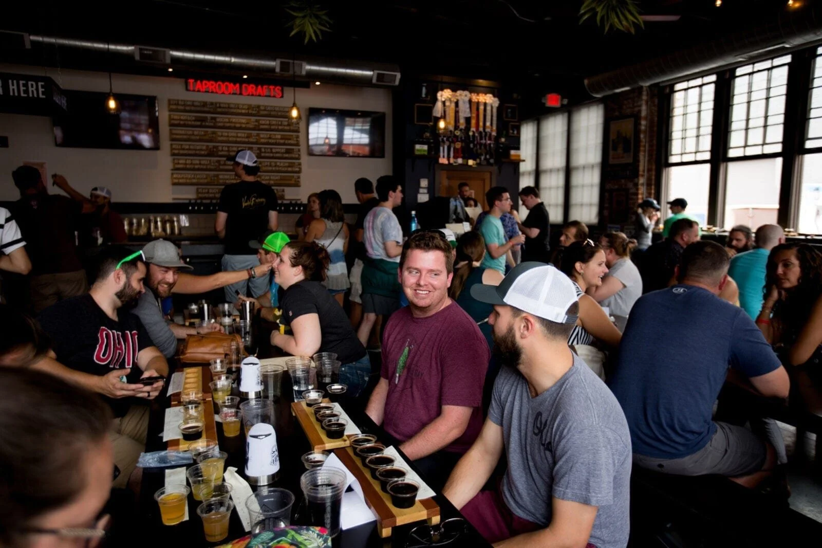 People sitting at a bar, participating in a beer tasting event, with some sharing drinks and enjoying the lively atmosphere of a brewery or pub.
