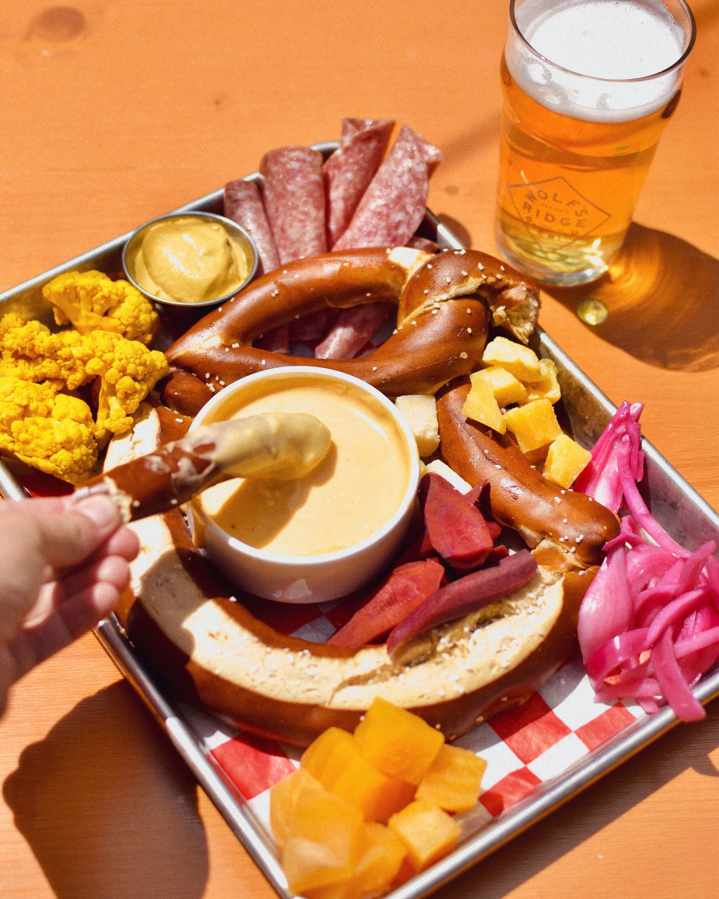 A tray of assorted snacks including pickled onions, cubes of cheddar cheese, sliced carrots, pretzels with cheese sauce, sliced salami, cauliflower florets with mustard, and a giant pretzel with cheese dip. A glass of beer is in the background.