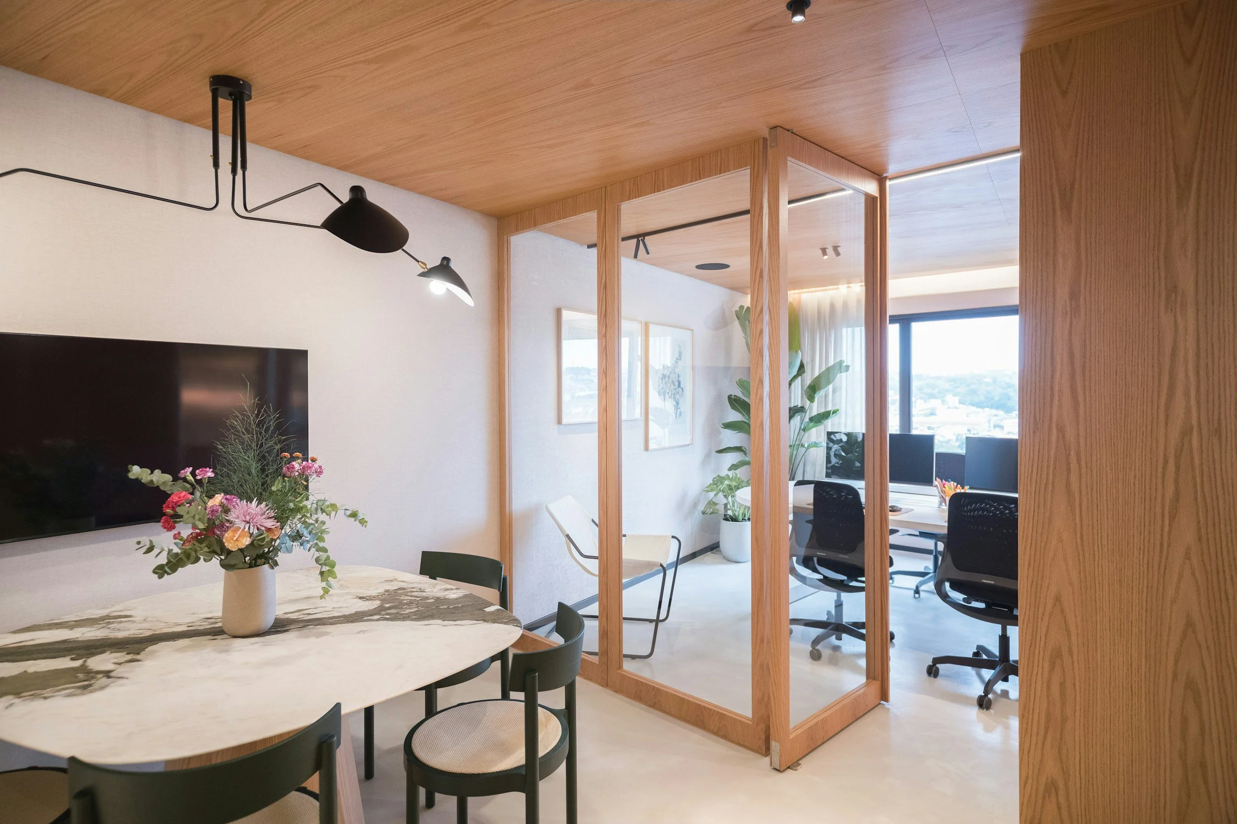 Modern office interior with wooden partition, wall art, potted plant, and a TV, with a view of city skyline through large window.