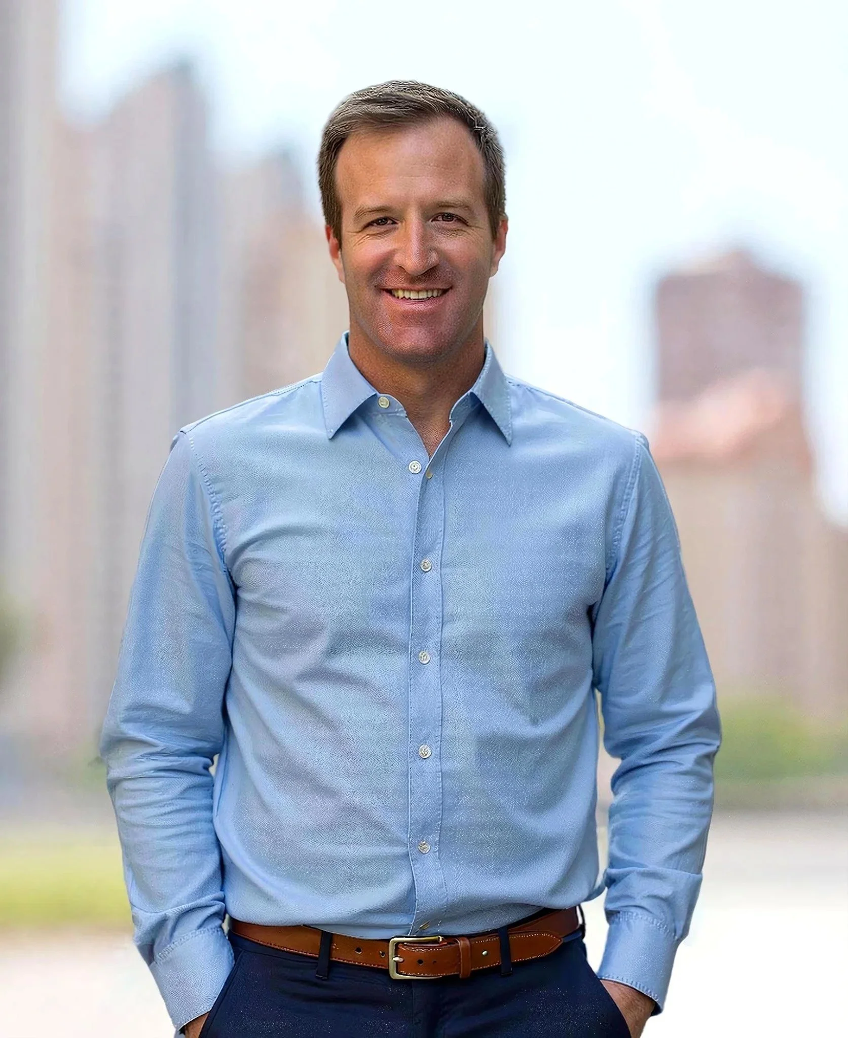 Portrait of Jordan Mullins in a light blue dress shirt with his hands in his pockets, standing outdoors with a blurred cityscape background.