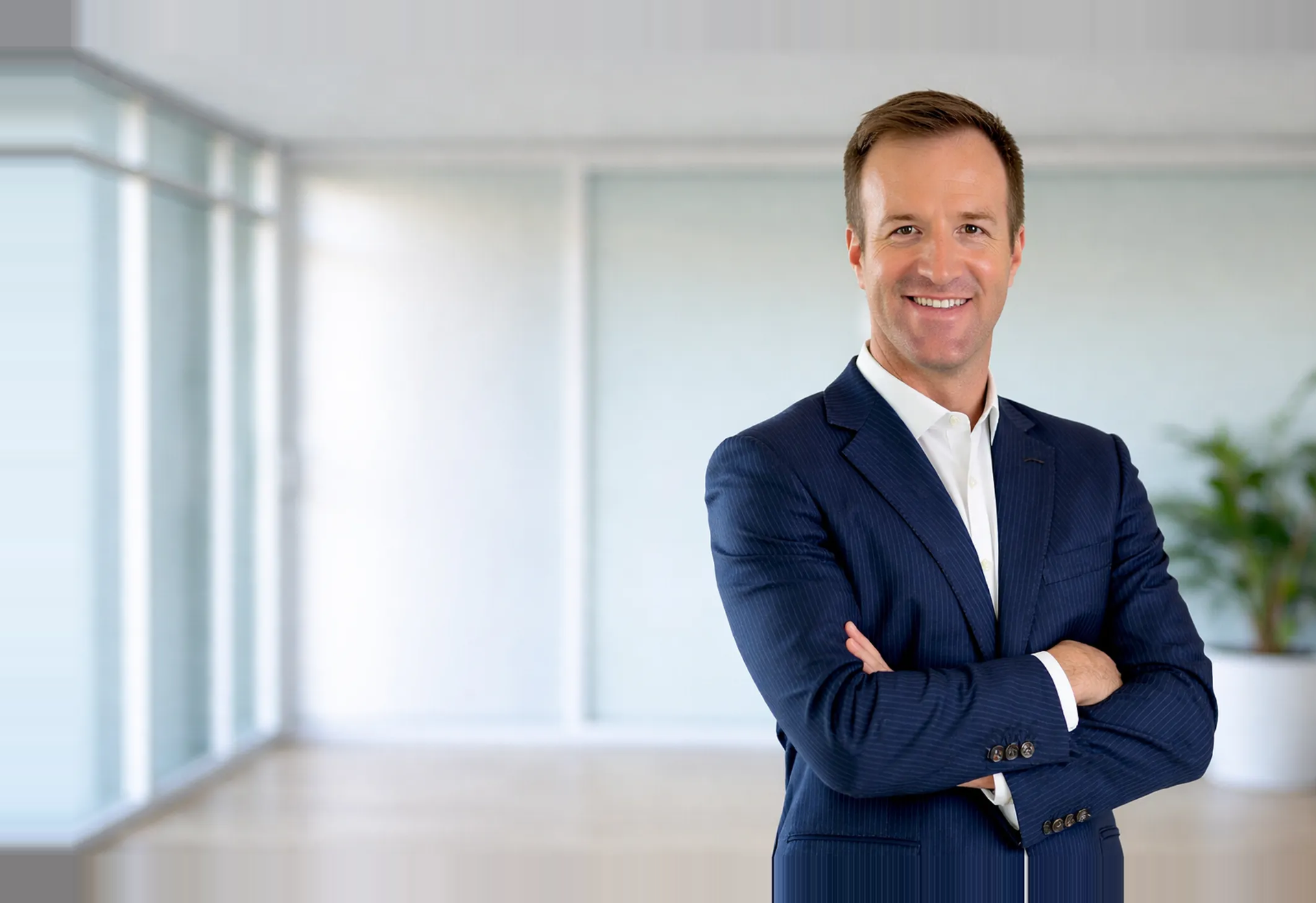 Austin Mediator Jordan Mullins, wearing a navy blue suit and white shirt standing with arms crossed in a conference room with large windows and a plant in the background.