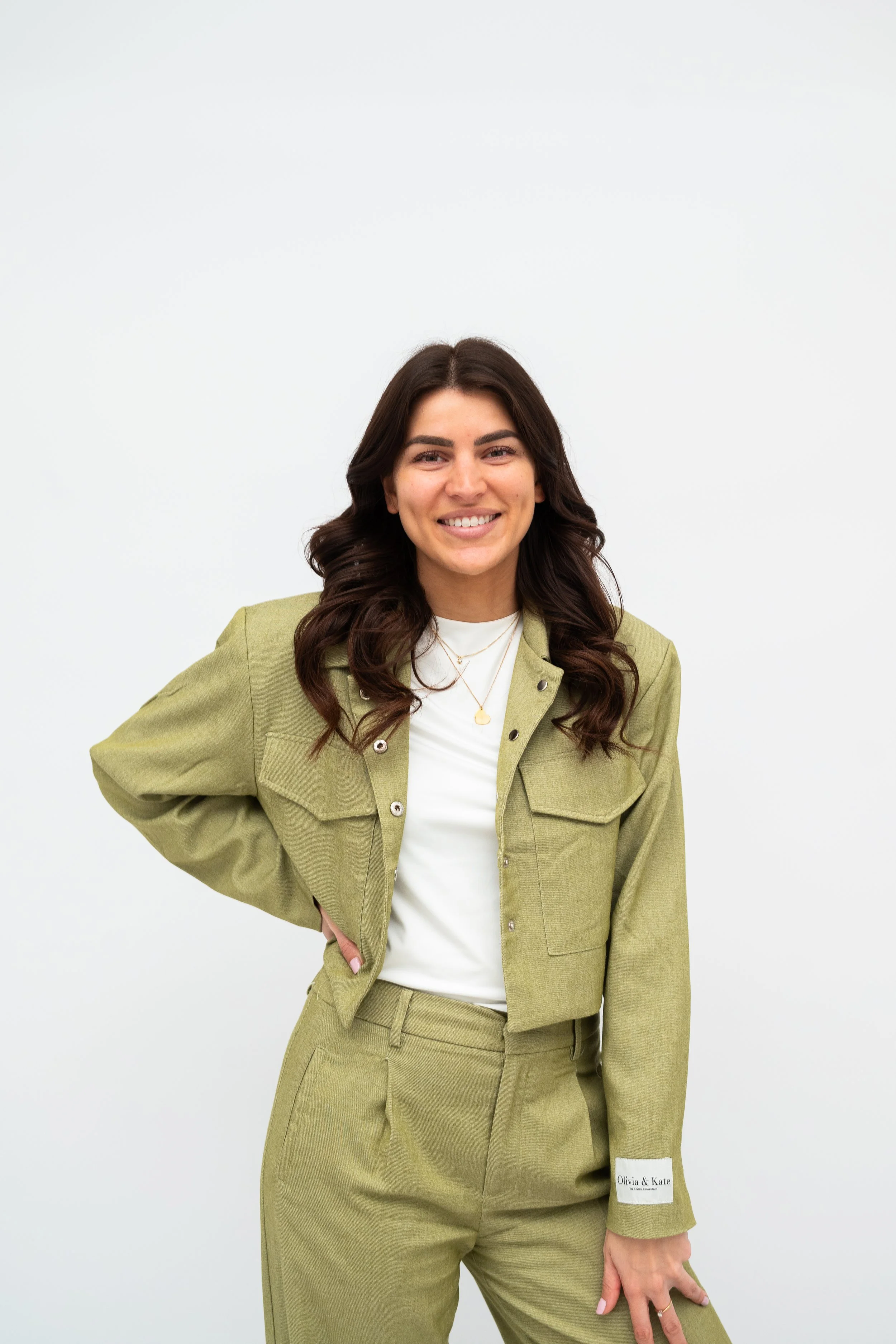 A woman with long dark hair, wearing a light green jacket and matching pants, smiling and standing against a plain white background.