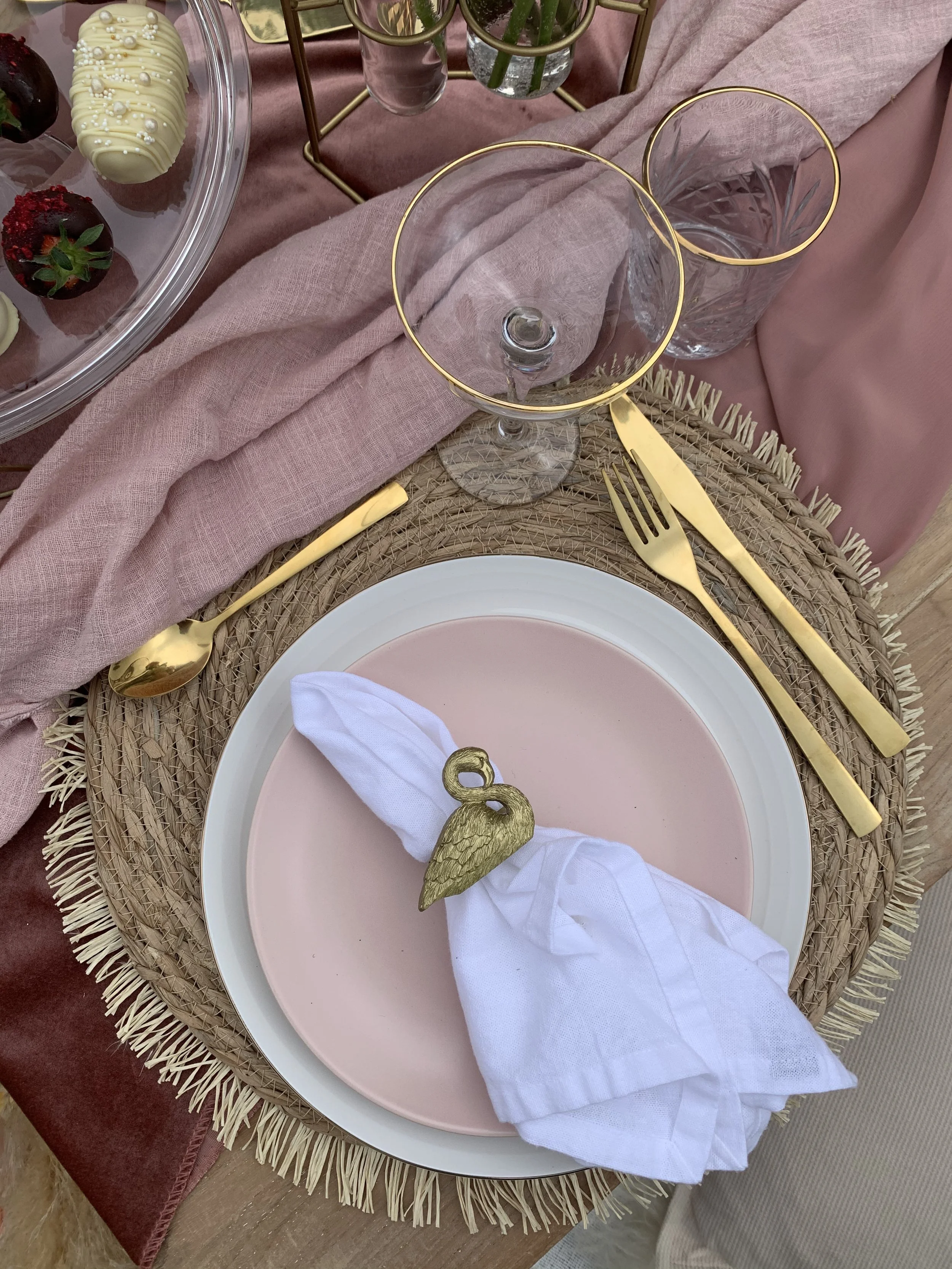 Table setting with pink and white plates, gold cutlery, clear glasses with gold rims, pink tablecloth, pink napkin with a gold swan napkin ring, and a partially visible tray of strawberries and chocolate-covered strawberries.