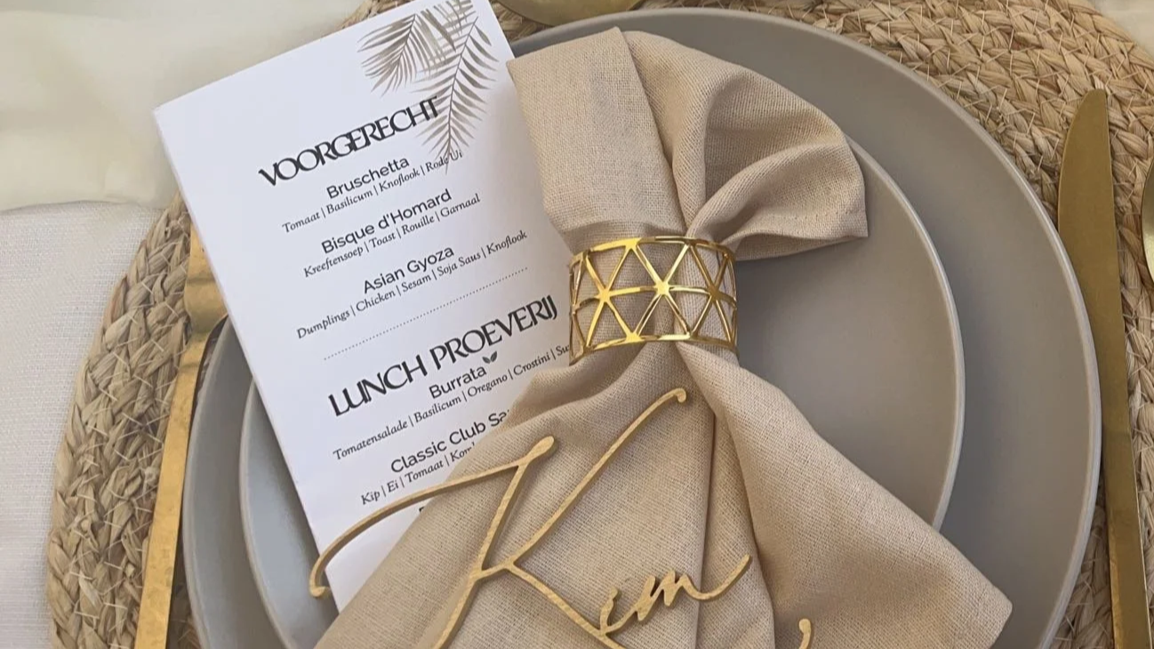 A dinner place setting with a beige napkin tied with a gold napkin ring, placed on a silver plate. A menu card is positioned on the napkin, listing Italian dishes. A gold-colored cutlery set is partially visible, with the handle of a fork and knife seen at the edge of the plate.