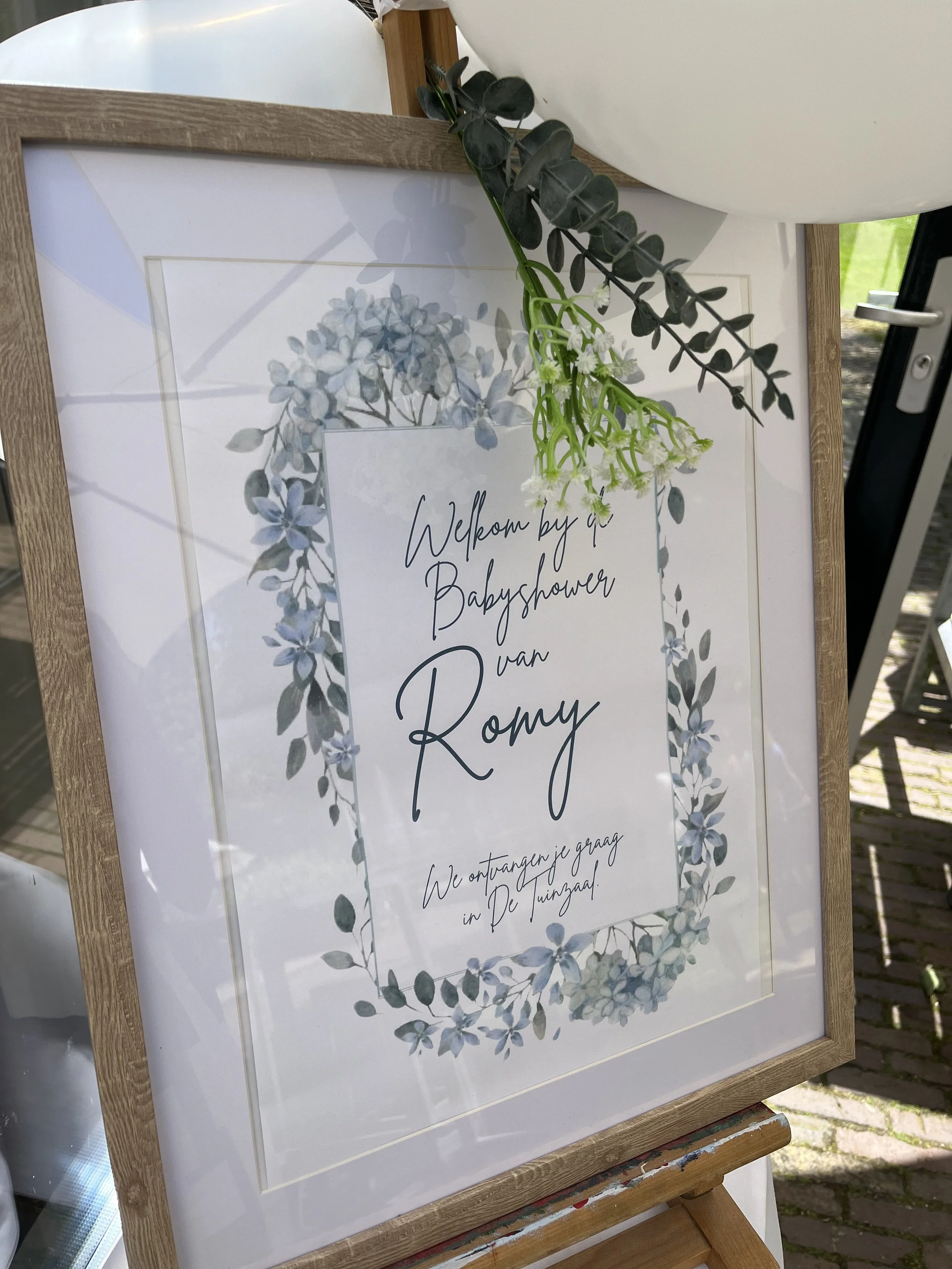 A framed sign with floral decorations welcoming guests to a baby shower for Romy, written in Dutch. A sprig of greenery is hanging from the top of the frame, and the sign is outdoors with sunlight and a cobblestone path visible.