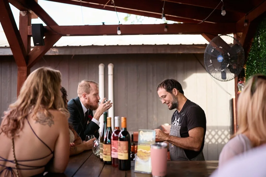 A man in a black shirt working behind a bar with bottles of wine on the counter, while a group of people sit around the bar, including a woman with red hair and another with blonde hair, outdoors under a wooden roof with hanging string lights.