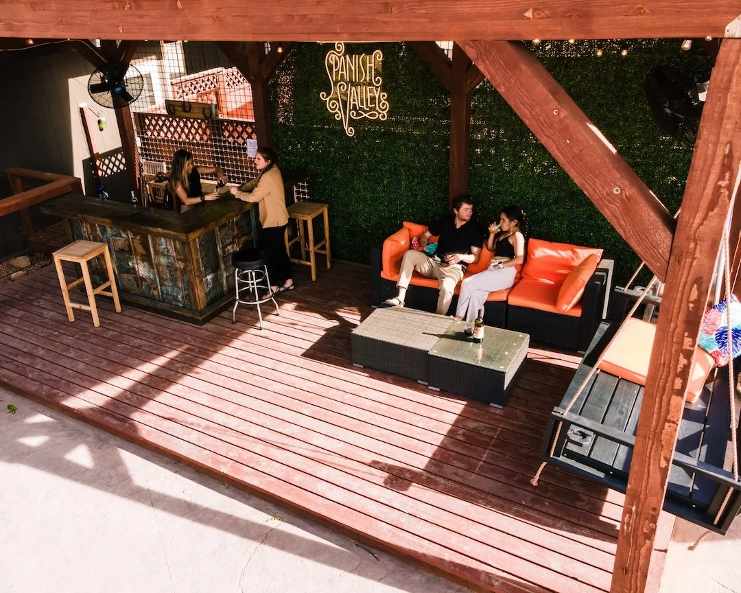 An outdoor patio area with a wooden deck, a bar on the left with two women talking, and a sitting area on the right with a man and a woman drinking beverages on a black and orange couch. There is a sign on the wall that reads 'Panshe Valley' and a la