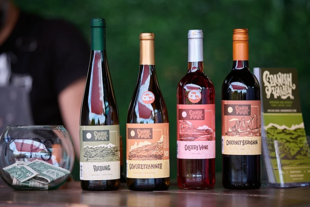 Four bottles of wine, a fishbowl with dollar bills, and a tourist map of Spanish Fork, Utah, on a wooden table outdoors.