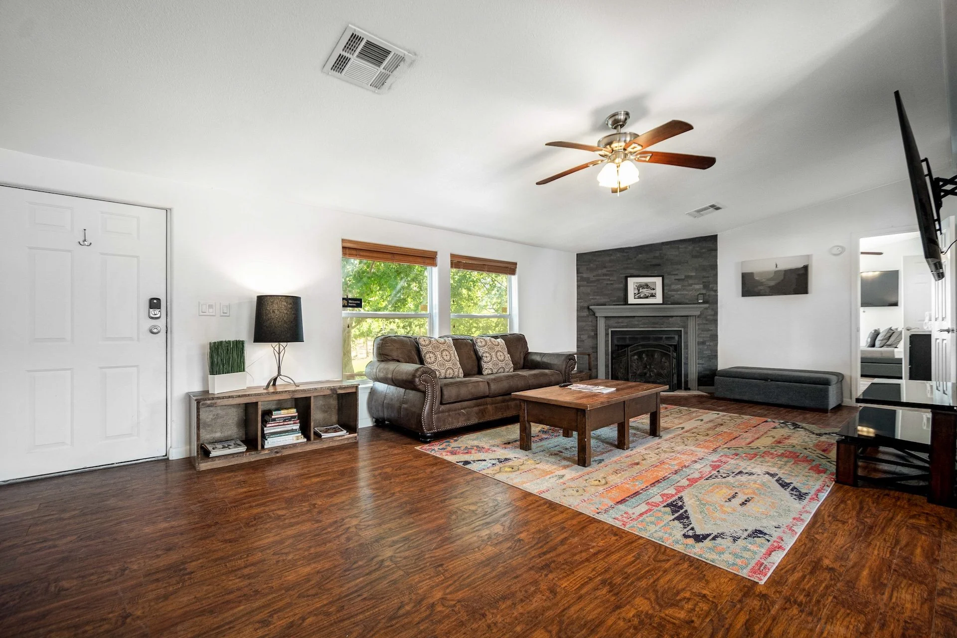 Living room with hardwood floors, a leather sofa, a wooden coffee table, a colorful area rug, a fireplace with a stone surround, windows with blinds, a ceiling fan, a modern TV, and a small bench, decor includes framed pictures and small plants.