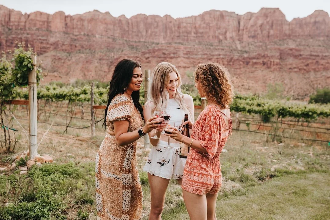 Three women standing outdoors in a vineyard with red rock formations in the background, holding glasses of red wine and smiling.