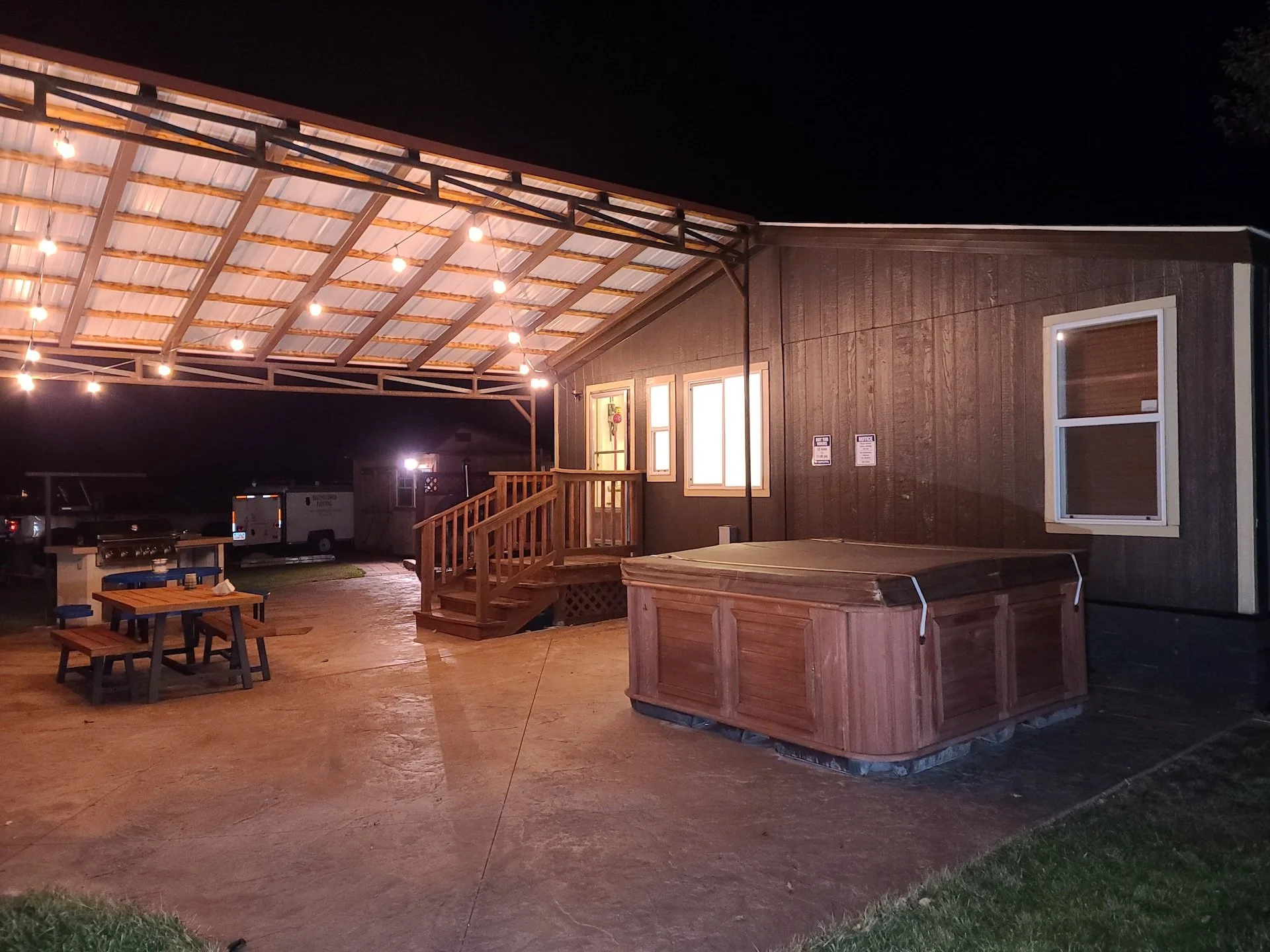 A covered outdoor space with string lights, wooden stairs leading up to a building, picnic tables, a hot tub, and parked vehicles at night.