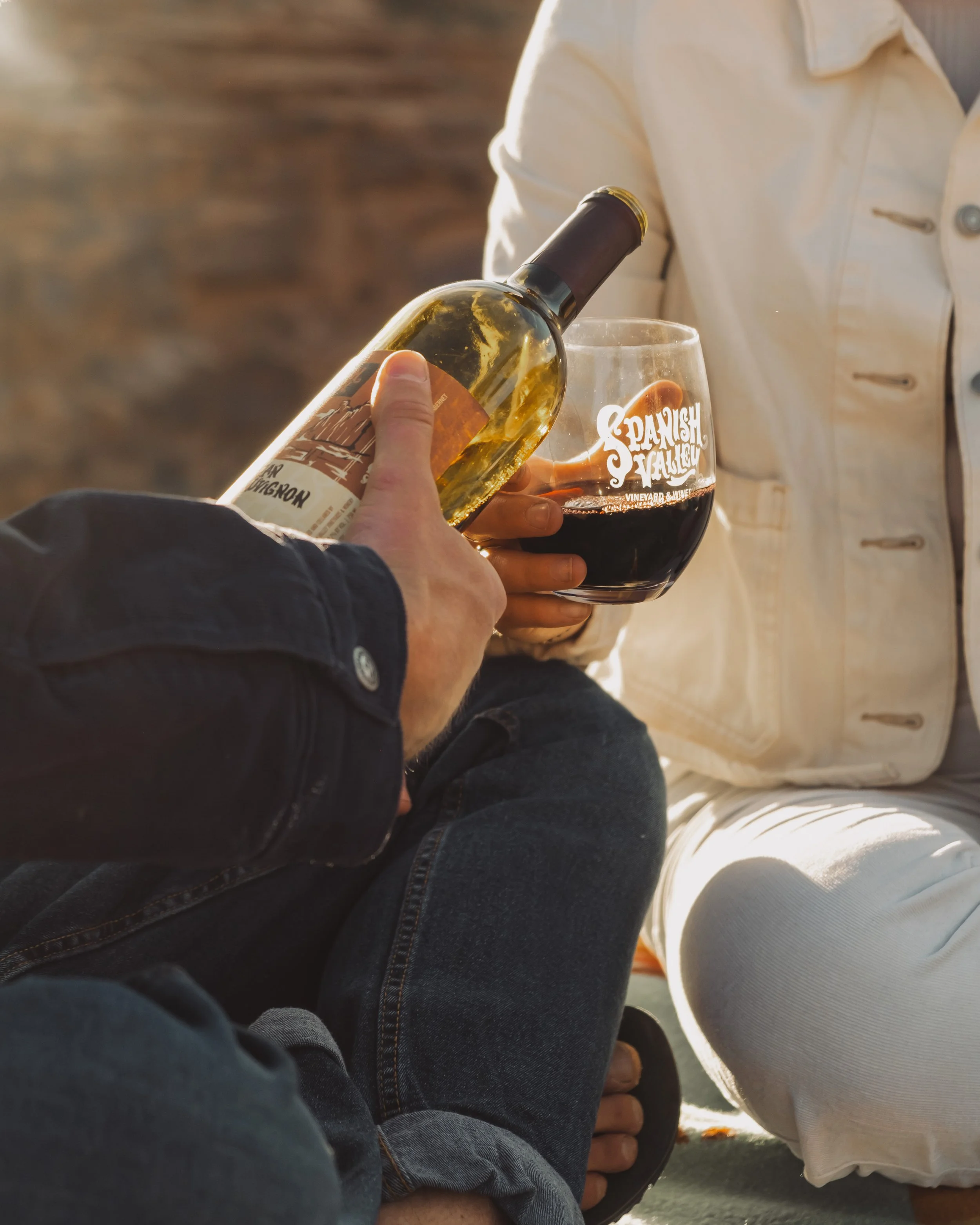 People sitting outside, one holding a glass of red wine, and another pouring white wine from a bottle.