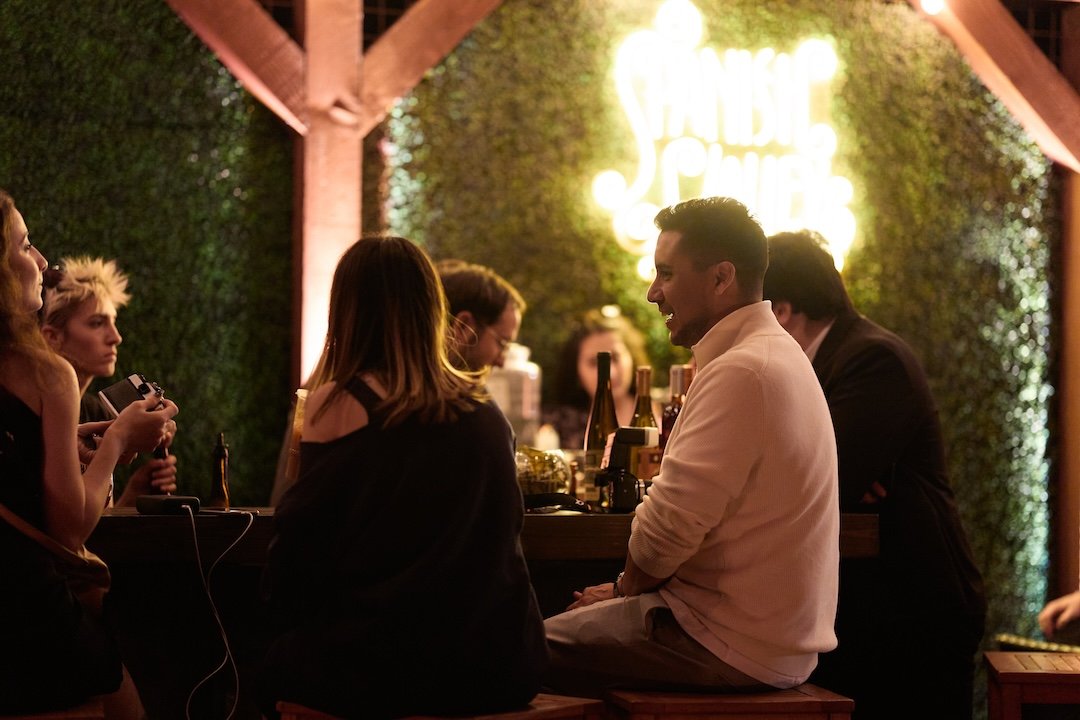 People socializing at a bar with a illuminated neon sign in the background, warm lighting, and a green leafy wall.