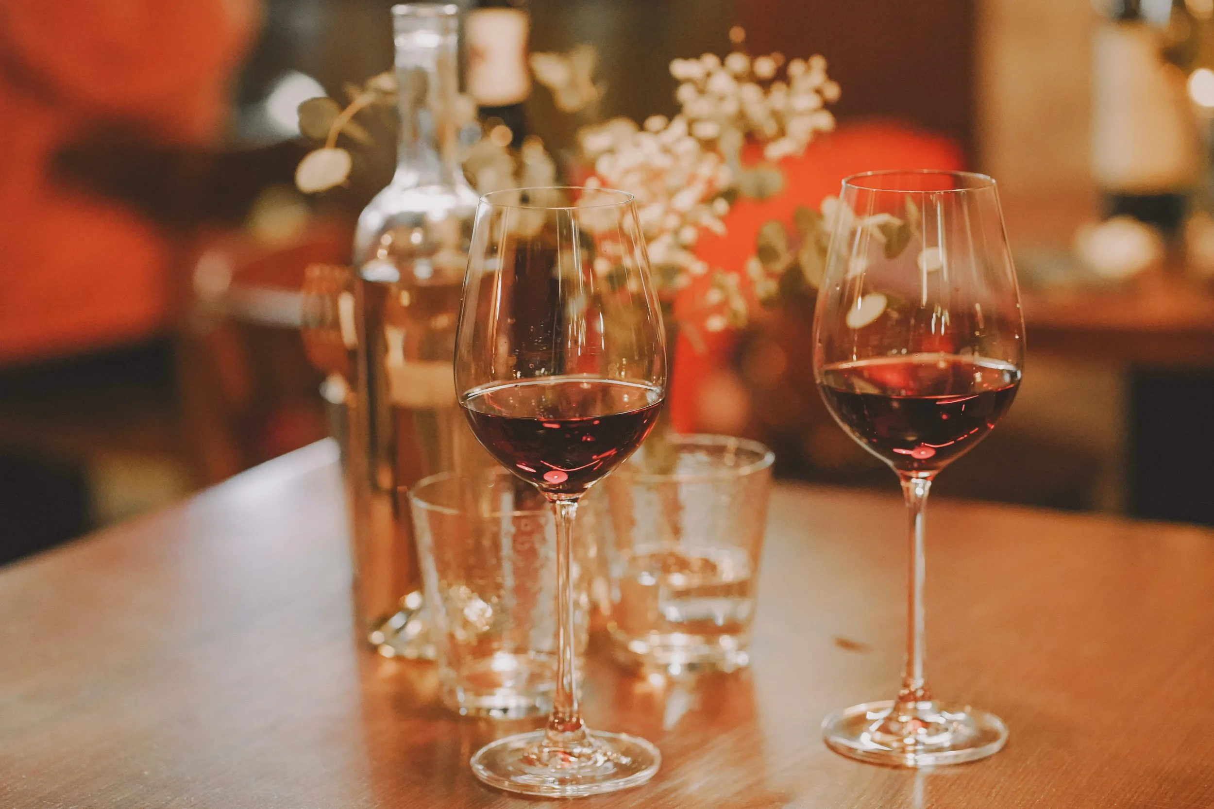 Two glasses of red wine on a wooden table with a blurry background of flowers and bottles.