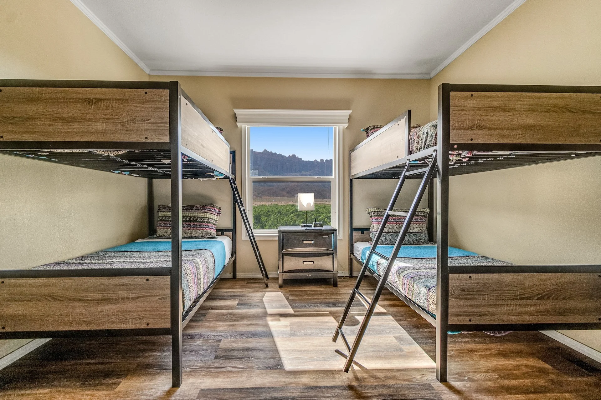 A room with two sets of bunk beds, a small black bedside table, two pillows on each bed, a window with a scenic view of mountains and greenery, and a table lamp on the table.
