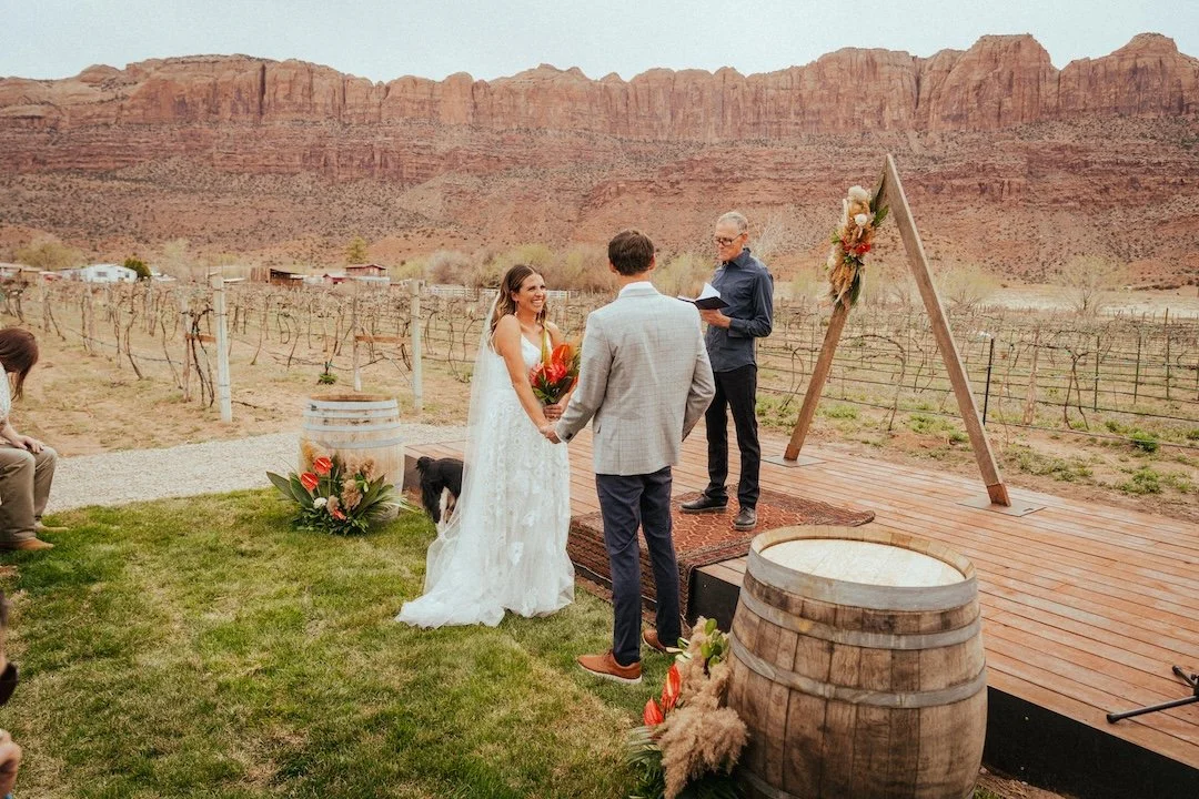 A wedding ceremony taking place outdoors in a desert landscape with red rock formations in the background. The bride and groom are holding hands in front of an officiant, with a rustic wooden arch decorated with flowers nearby. There are wine barrels