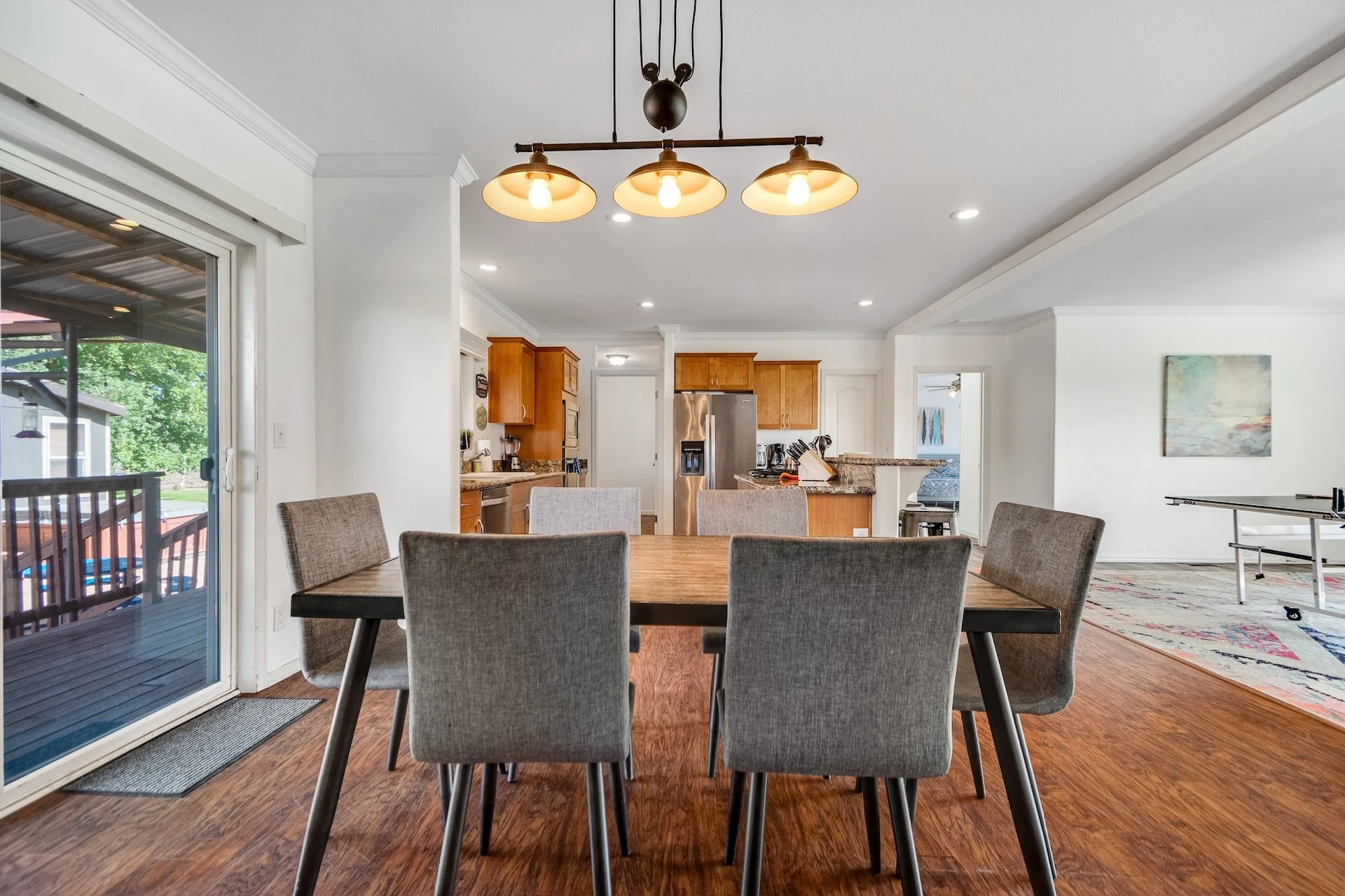 Dining area with a wooden table and six gray upholstered chairs, sliding glass door leading to a deck, view of the kitchen with wooden cabinets and stainless steel appliances, pendant light fixture above the table, hardwood floor, and a living room w