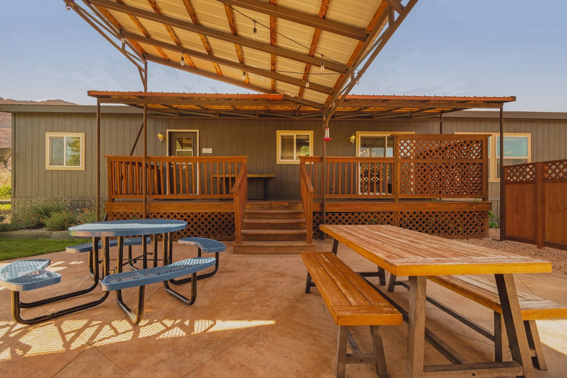 A backyard patio with a wooden deck, picnic tables, and a shaded area with string lights. The deck is attached to a gray house with windows, and a wooden staircase leads up to the deck. The patio has a concrete floor and a garden with plants on the side.