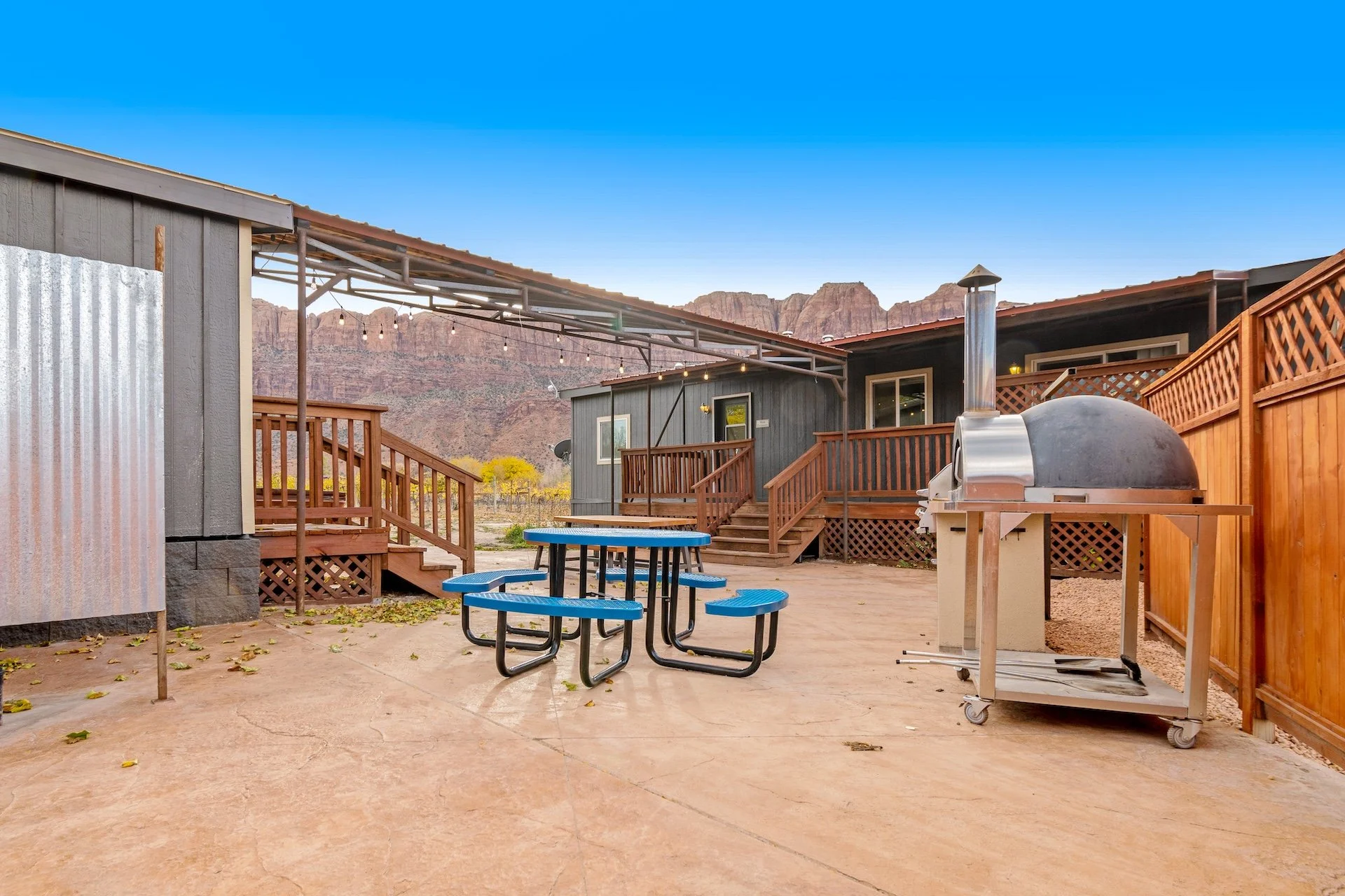 A backyard patio area with a blue picnic table, a stainless steel pizza oven, and a wooden deck with stairs, surrounded by a brown wooden fence, with mountains in the background and a clear blue sky.