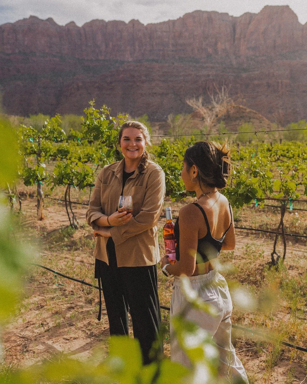 Two women standing in a vineyard, one smiling and holding a glass of wine, the other holding a bottle of rosé, with mountains in the background during daytime.