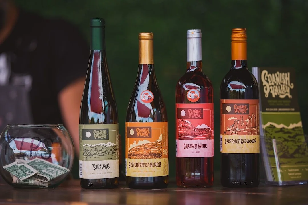 Four bottles of wine with colorful labels, a fishbowl with money, and a leaf-green informational stand on a wooden surface.