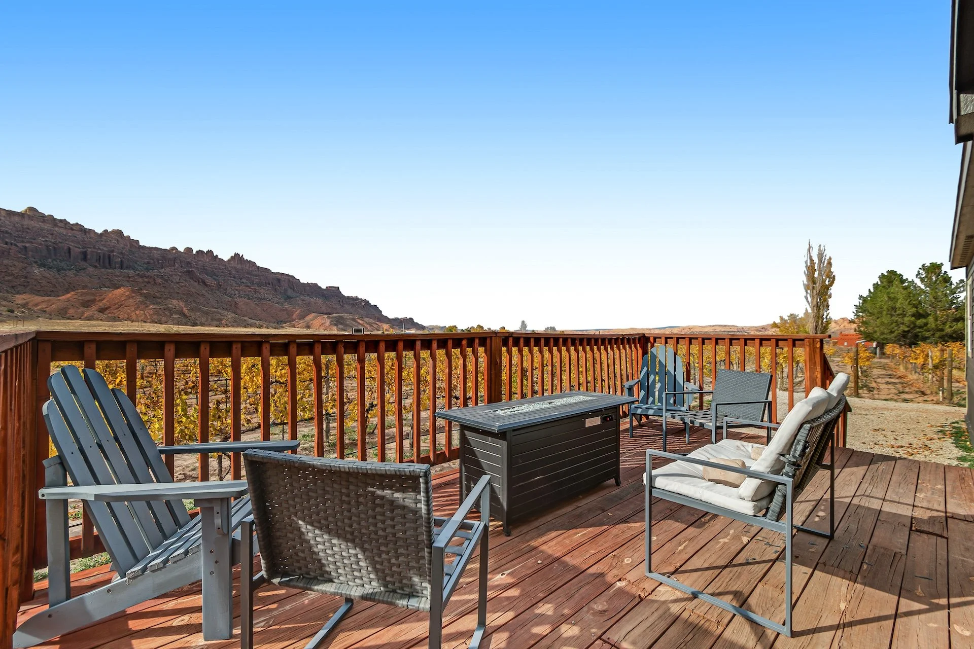A wooden deck with outdoor furniture including chairs and a fire pit, overlooking a desert landscape with mountains and trees during daytime.