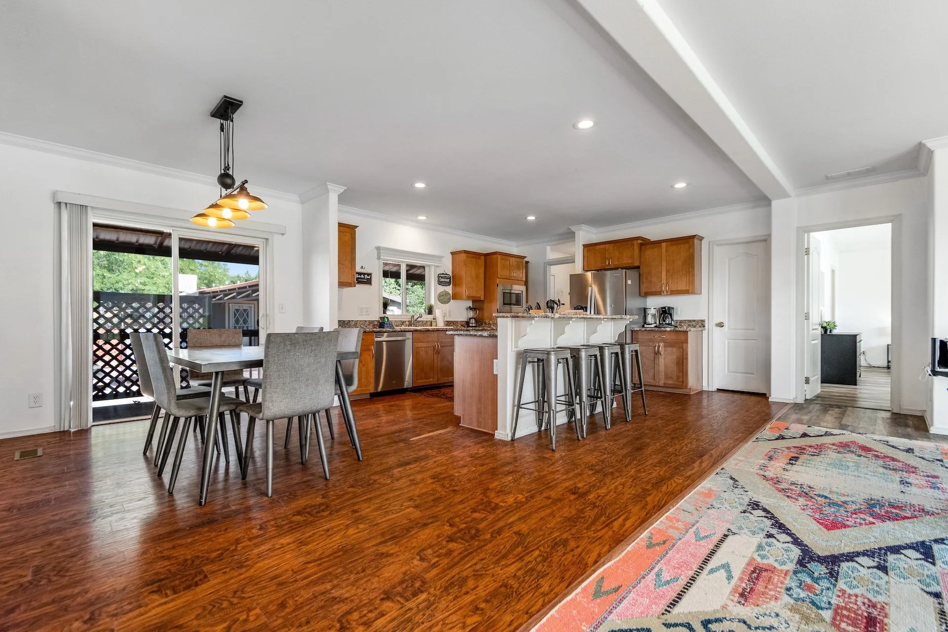 Open-concept kitchen and dining area in a home with wooden floors, white walls, and natural light from a sliding glass door and windows. The kitchen features wooden cabinets, stainless steel appliances, a granite countertop, and a small breakfast bar