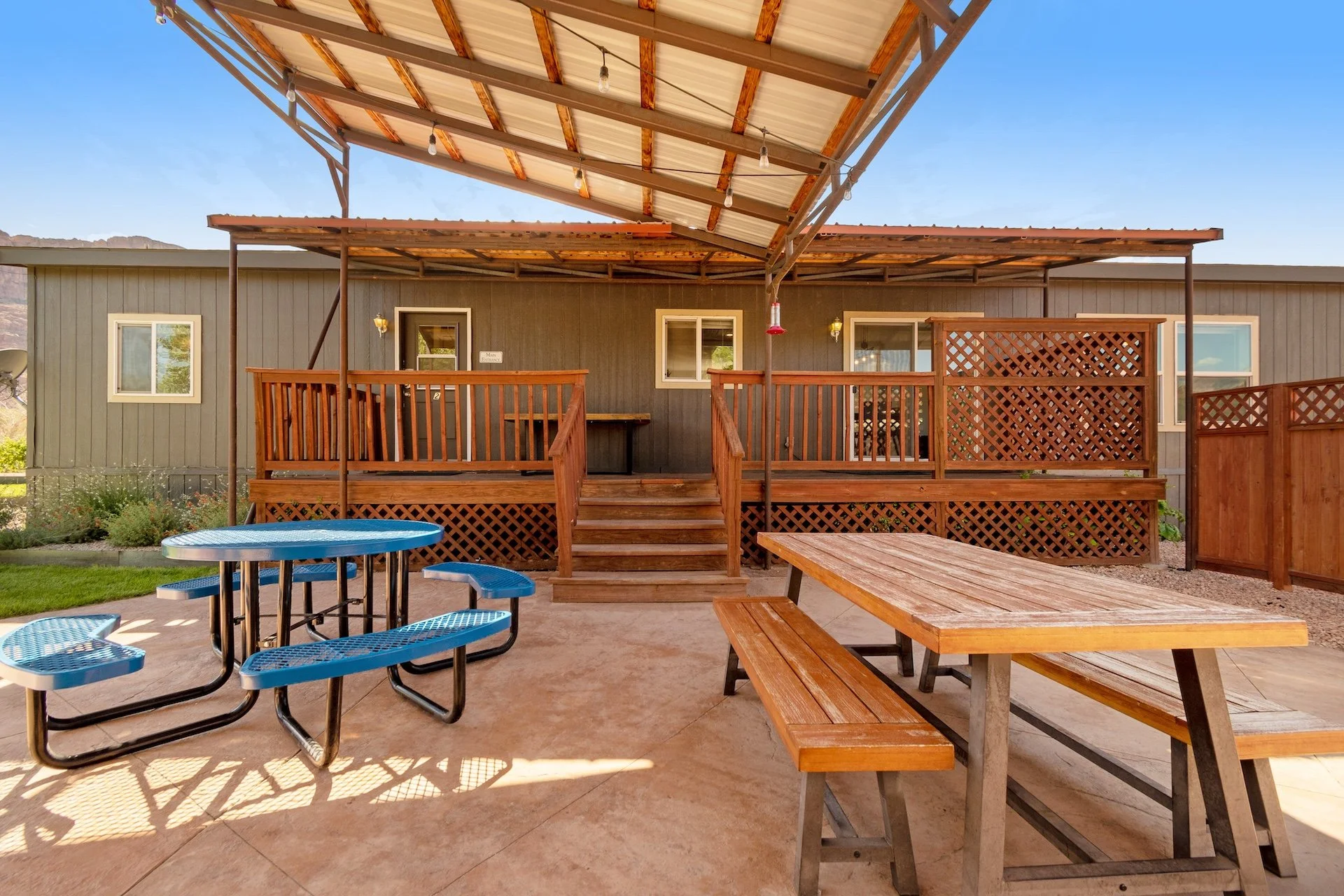 An outdoor patio area with a wooden deck and a sloped metal roof. There are steps leading up to the deck, which is attached to a gray building with white-framed windows and doors. The patio features a wooden picnic table with benches, a blue metal pi
